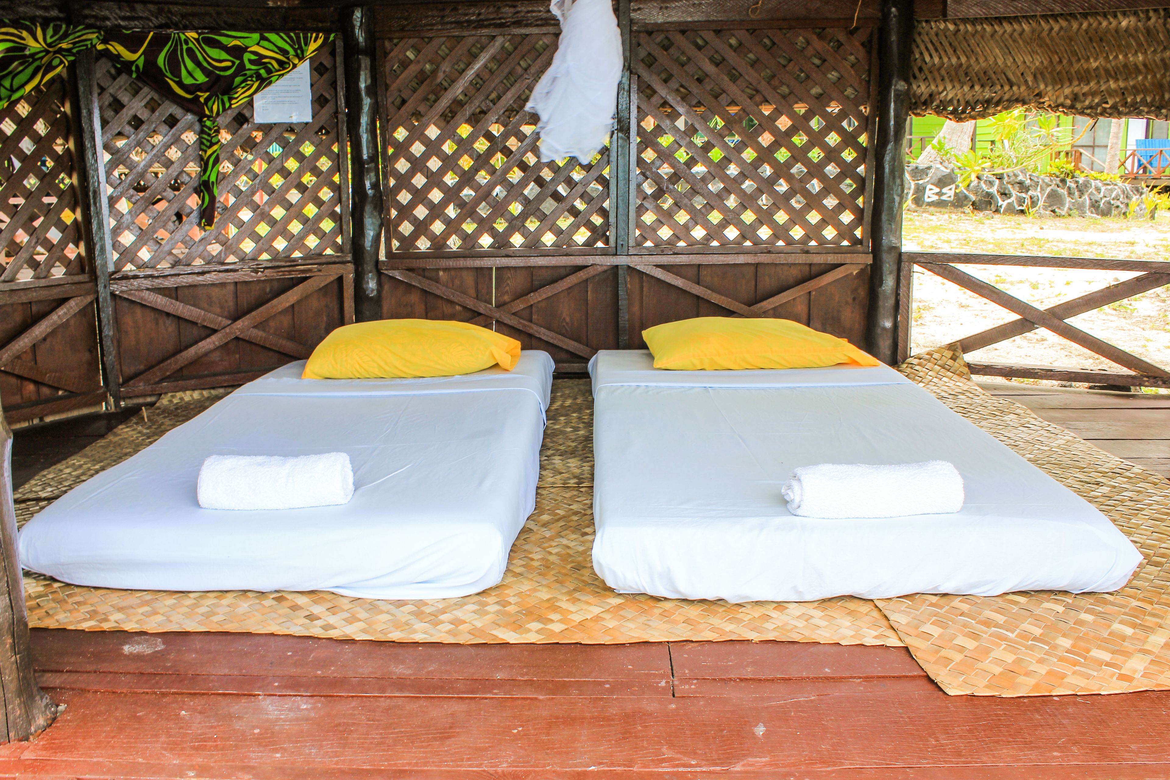 Two single beds with white and yellow pillows, a woven mat, and a wooden structure with a lattice design.