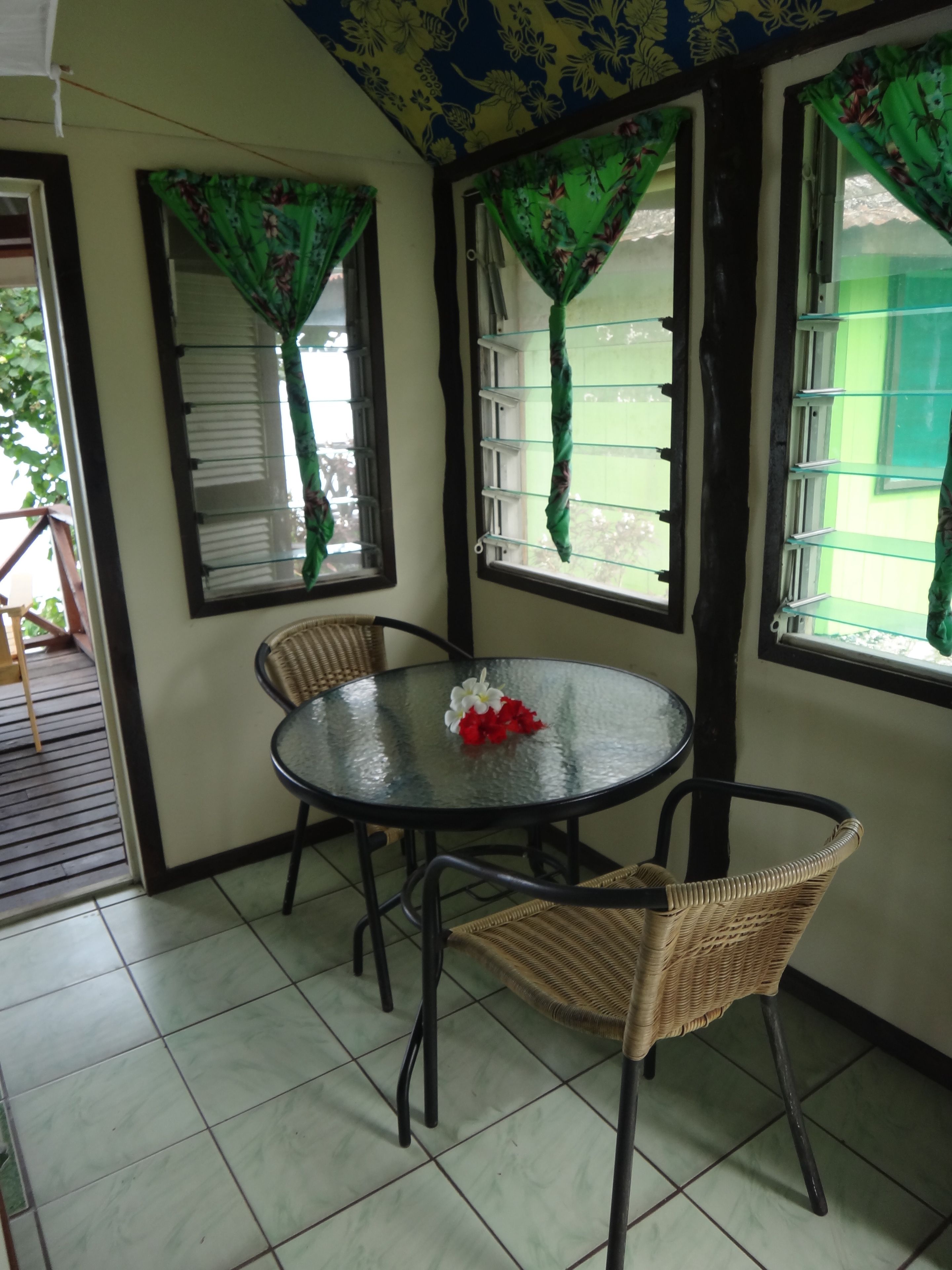 A small, enclosed patio area with a round glass table and two wicker chairs. The walls are painted light green, and there are windows with green floral curtains.