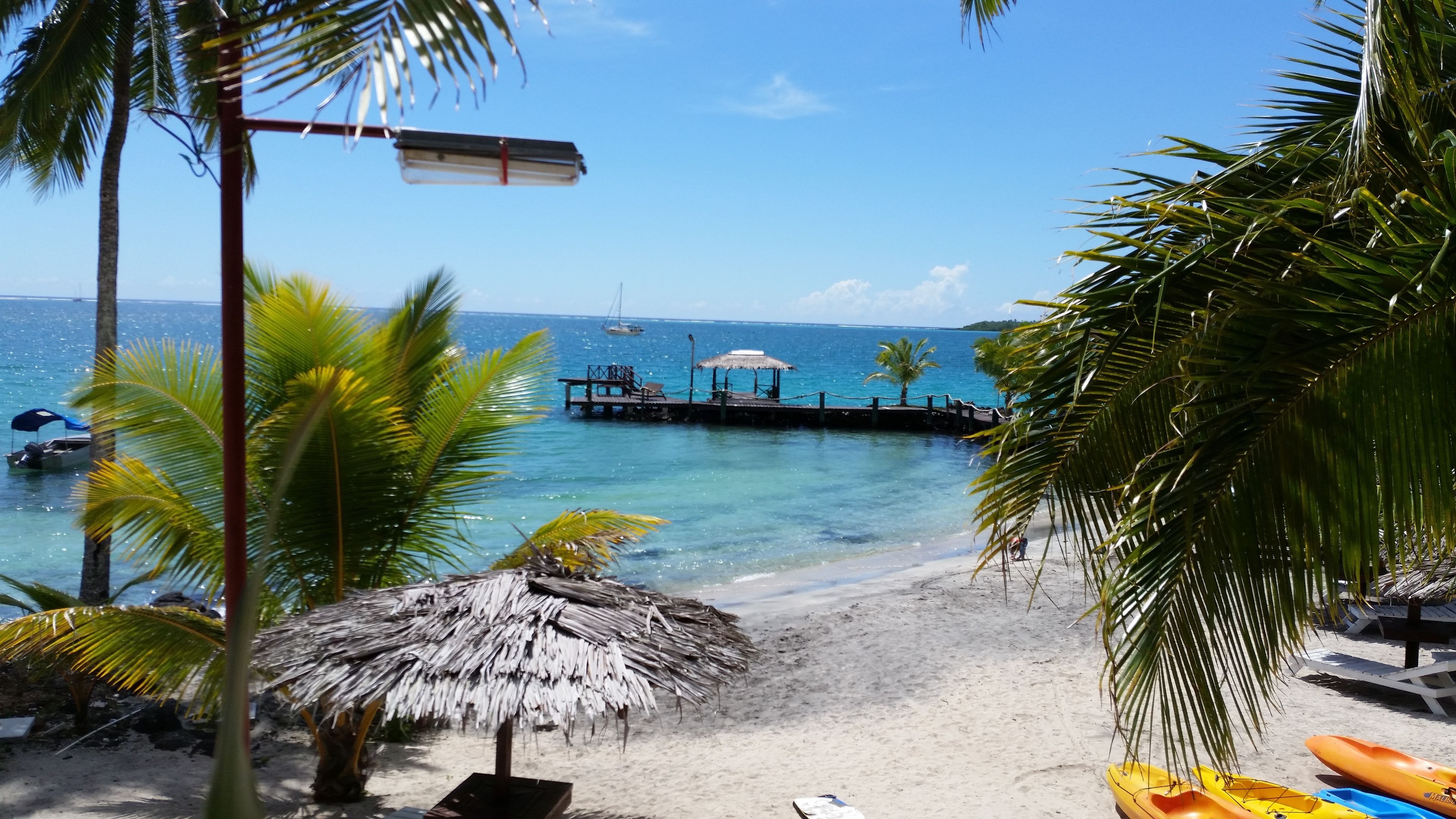 A beach with a thatched umbrella, kayaks, and a pier extending into the ocean.