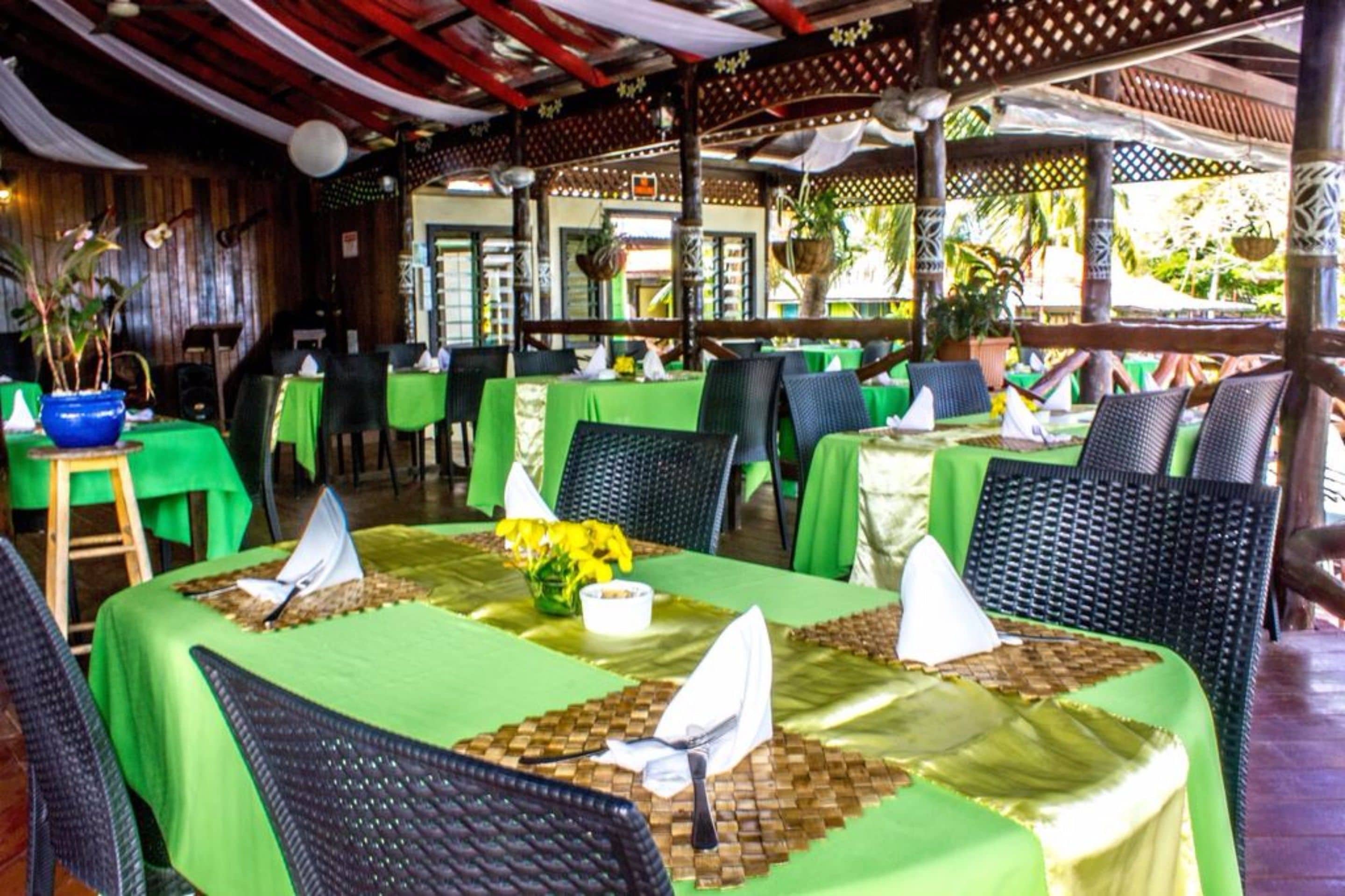 A restaurant with green tablecloths, wicker chairs, and a tropical setting with plants and a wooden ceiling.