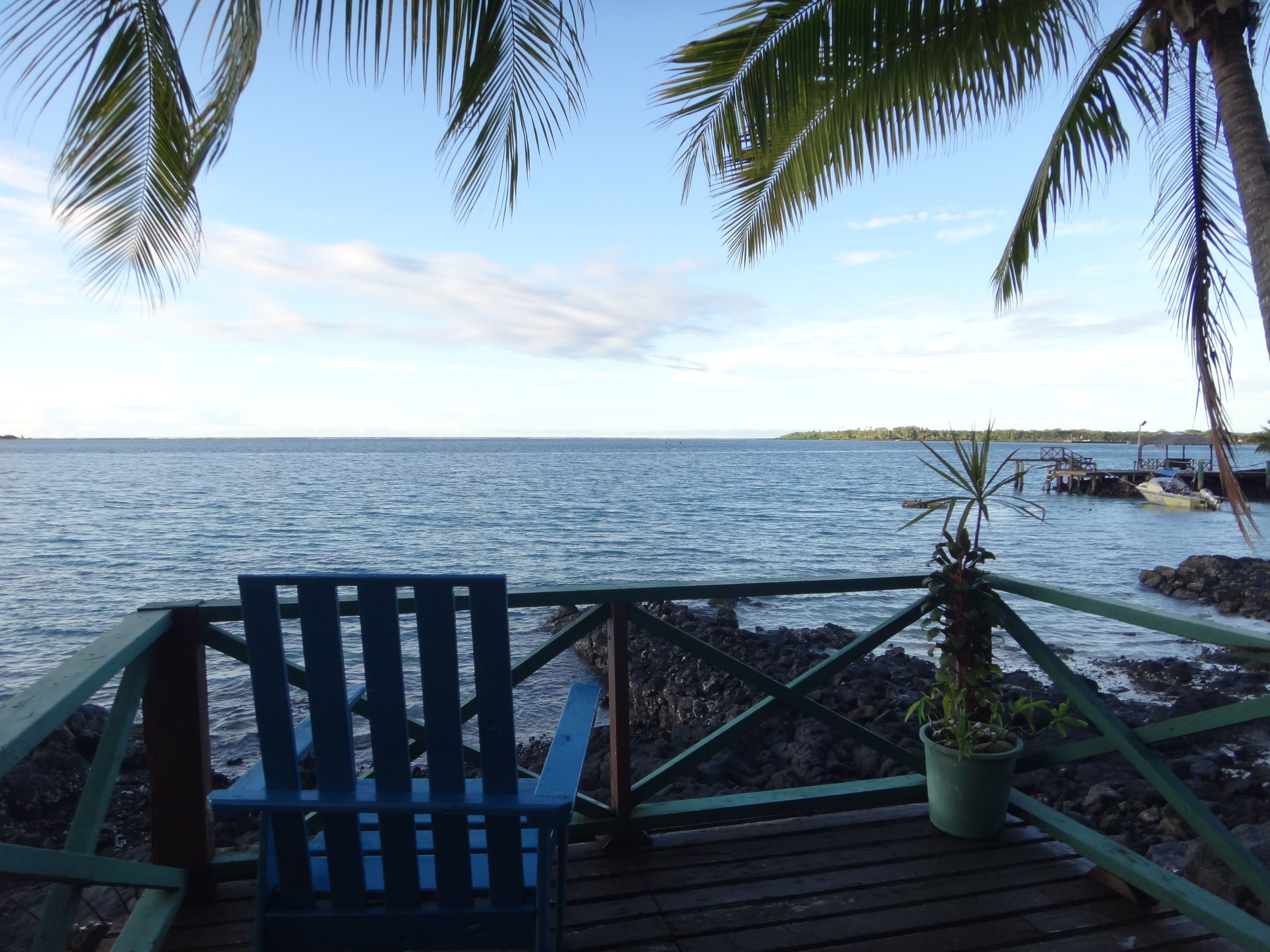 Una terraza de madera con una silla azul y una planta en maceta, con vista a un mar tranquilo y una isla distante.