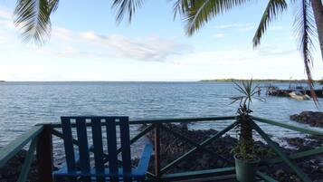 Una terraza de madera con una silla azul y una planta en maceta, con vista a un mar tranquilo y una isla distante.