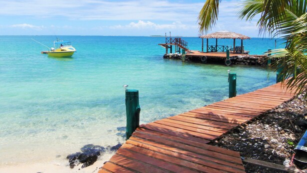 A wooden dock extending into clear turquoise water, a small boat anchored nearby, and a thatched-roof structure with seating. - Va-i-moana Seaside Lodge (Asau)