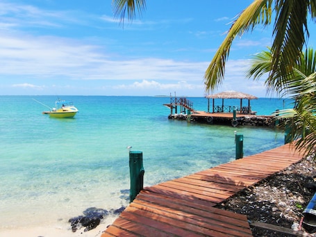 A wooden dock extending into clear turquoise water, a small boat anchored nearby, and a thatched-roof structure with seating.