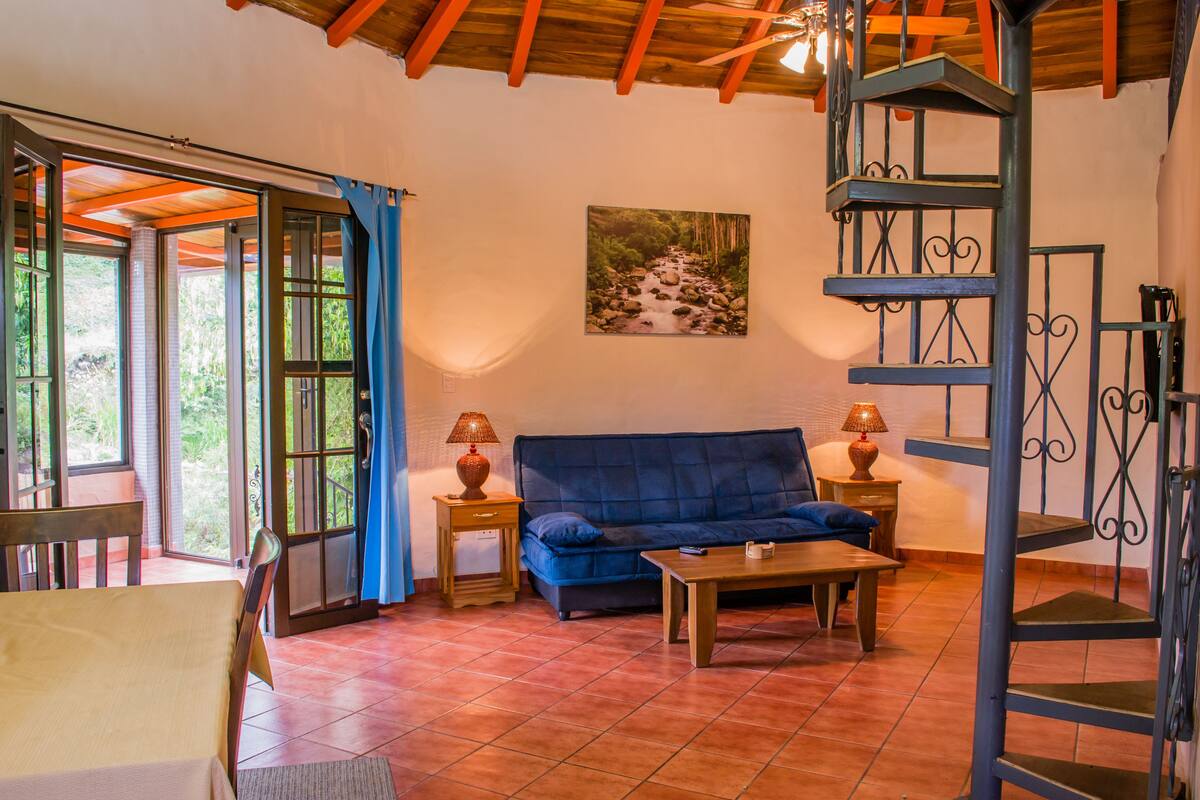 A living room with a blue sofa, wooden coffee table, and two side tables with lamps. There is a painting on the wall and a spiral staircase.