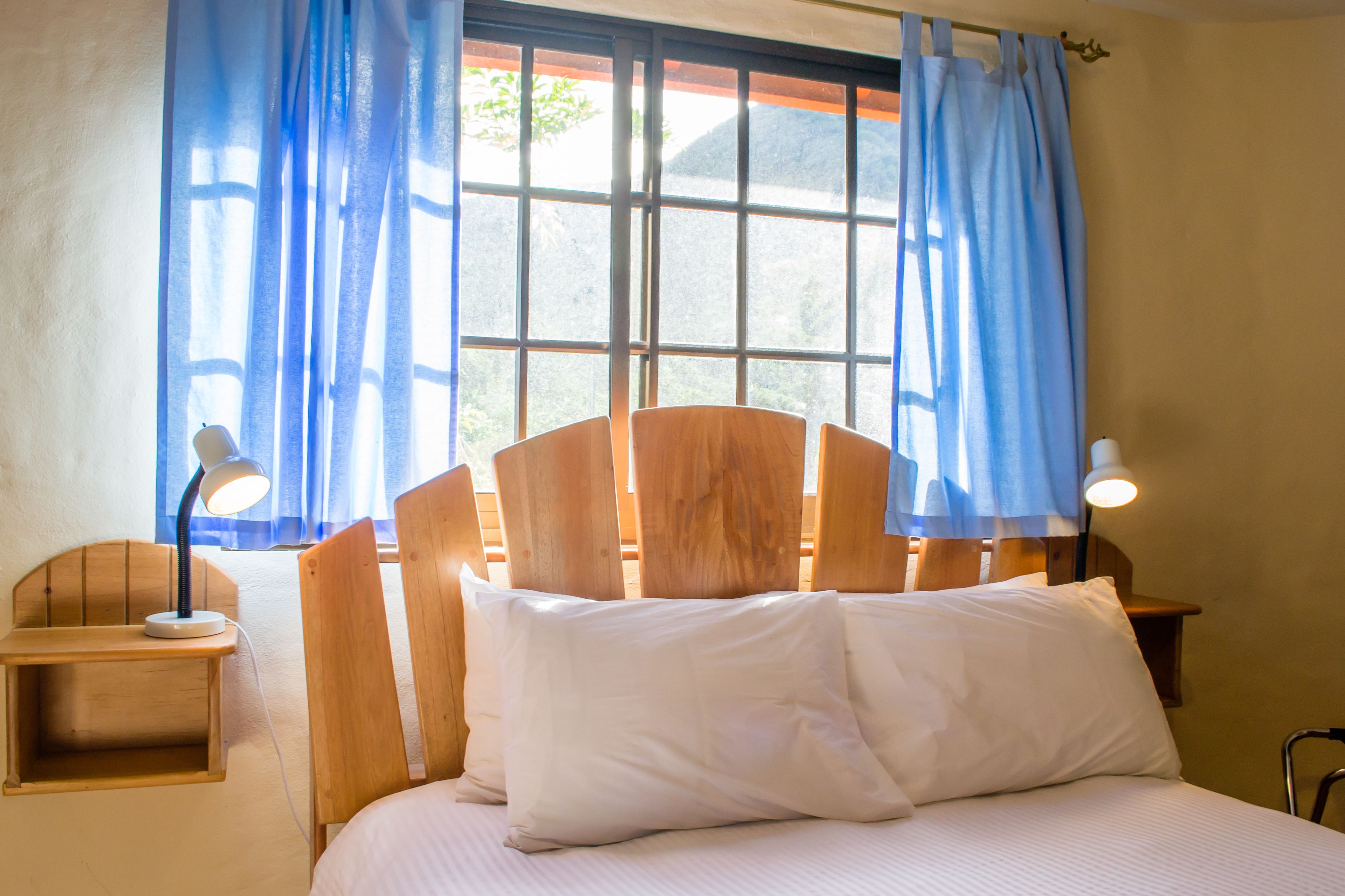 A bed with a wooden headboard, two bedside lamps, a nightstand, and a window with blue curtains.