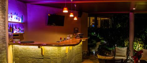 Uma área de bar com móveis de vime, um balcão com garrafas e uma televisão fixada na parede.