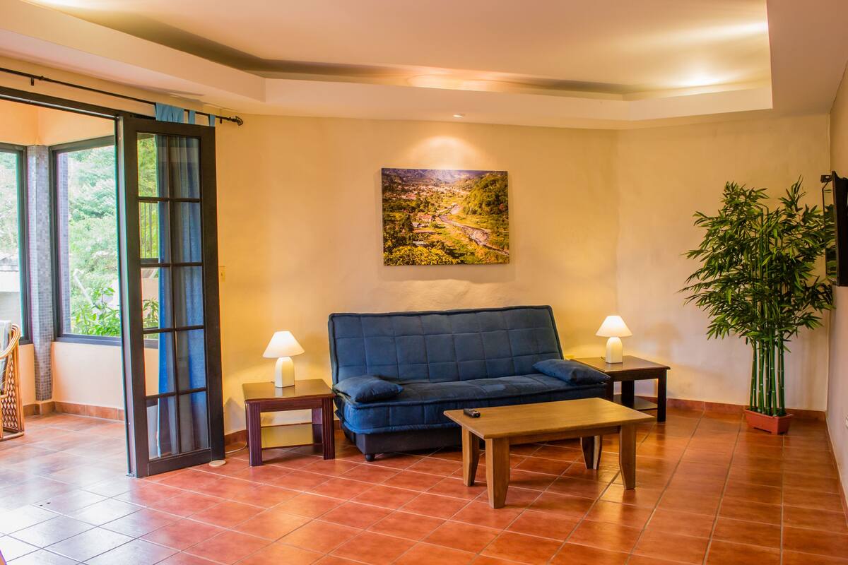 A living room with a blue sofa, two side tables with lamps, a wooden coffee table, a large painting on the wall, and a potted plant.
