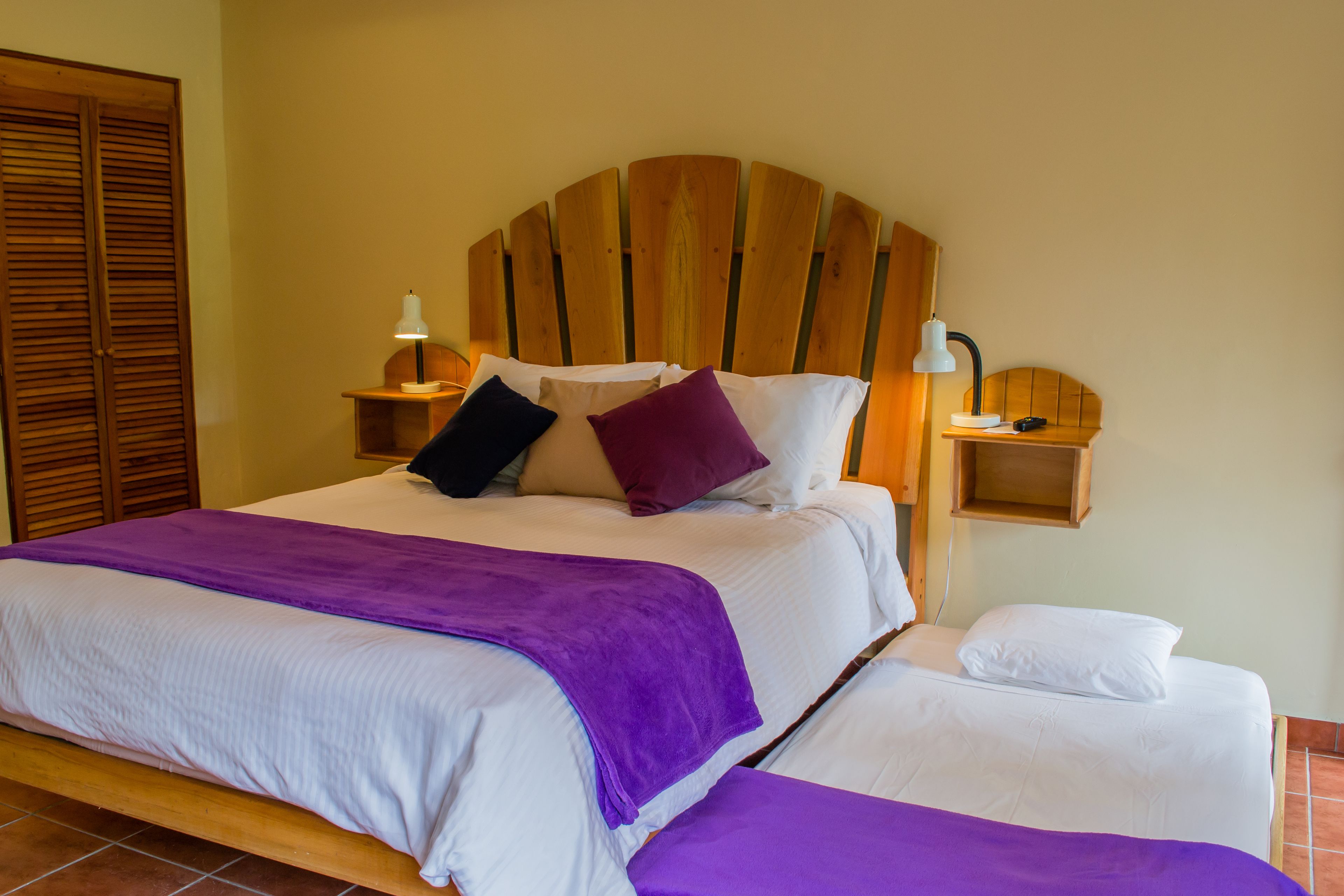 Un lit double avec une tête de lit en bois, deux tables de chevet avec des lampes et un plaid violet.