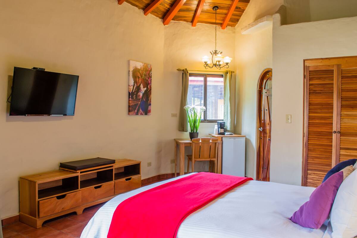 A bedroom with a bed, a TV, a desk, a chair, a window with curtains, and a wooden wardrobe.