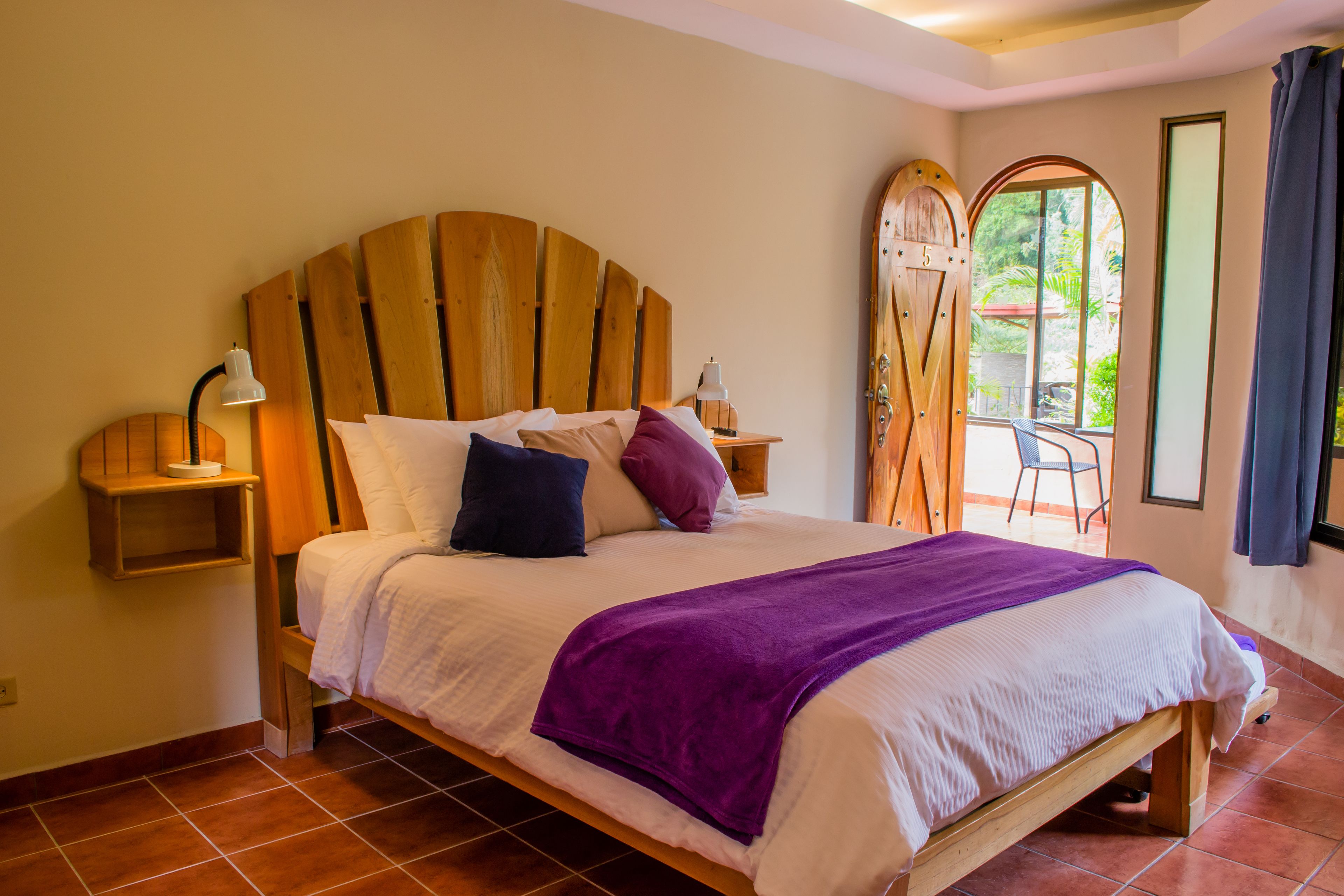 A bedroom with a wooden headboard, a bed with white and purple bedding, a purple throw blanket, a nightstand with a lamp, and a view of the outdoors through a door.