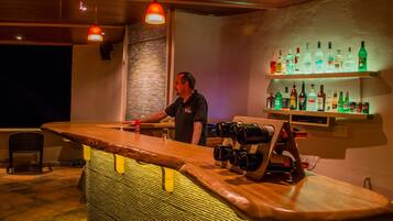 Una zona de bar con un cantinero, una barra de madera y estantes llenos de botellas de licor.