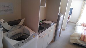 A compact hotel room with a washing machine, a refrigerator, a small kitchen area, and a bed with a striped blanket.