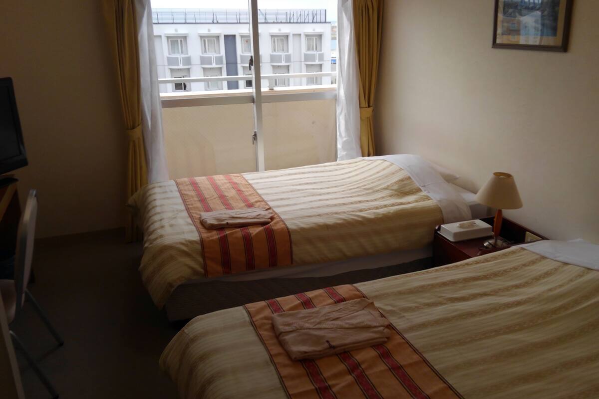 Une chambre d’hôtel avec deux lits, une télévision, une lampe de chevet et une fenêtre donnant sur des immeubles.