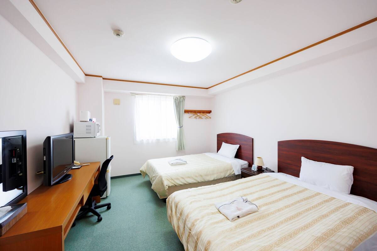 Ein Hotelzimmer mit zwei Betten, einem Schreibtisch mit Computer, einem Fernseher und einem Fenster mit Vorhängen.