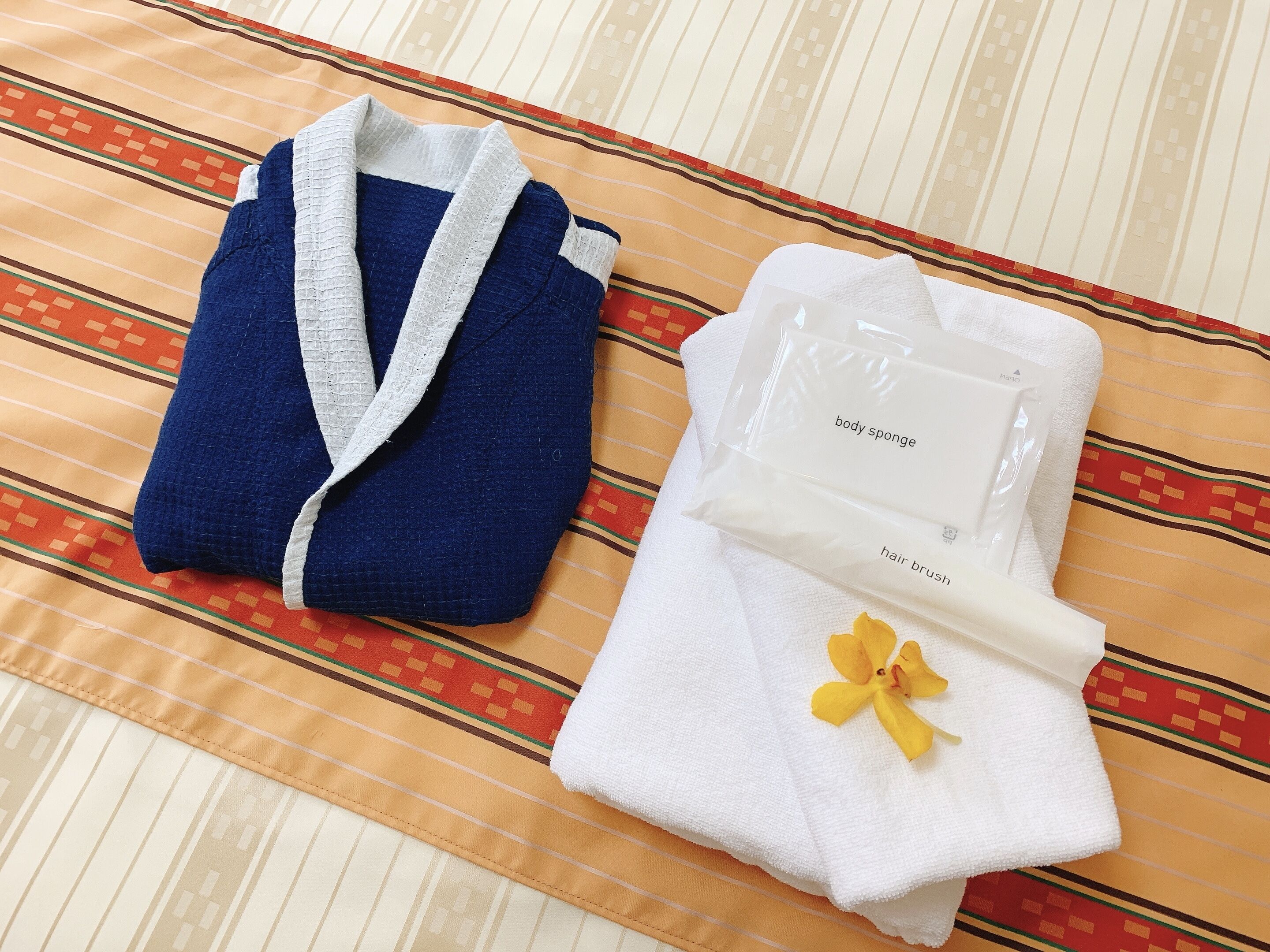 Un albornoz azul doblado con cuidado y una toalla blanca con un detalle floral amarillo, sobre un edredón a rayas.