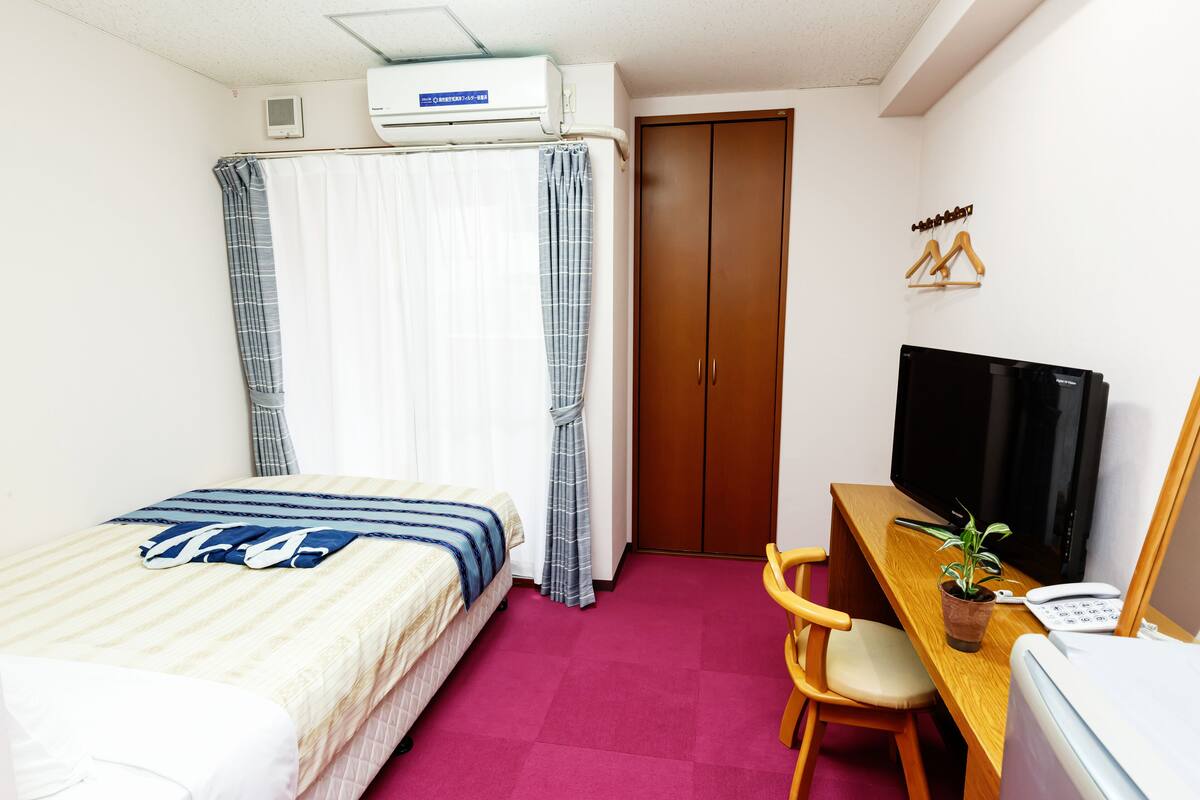 A hotel room with a bed, a desk with a computer and a plant, a wardrobe, and a shower curtain.