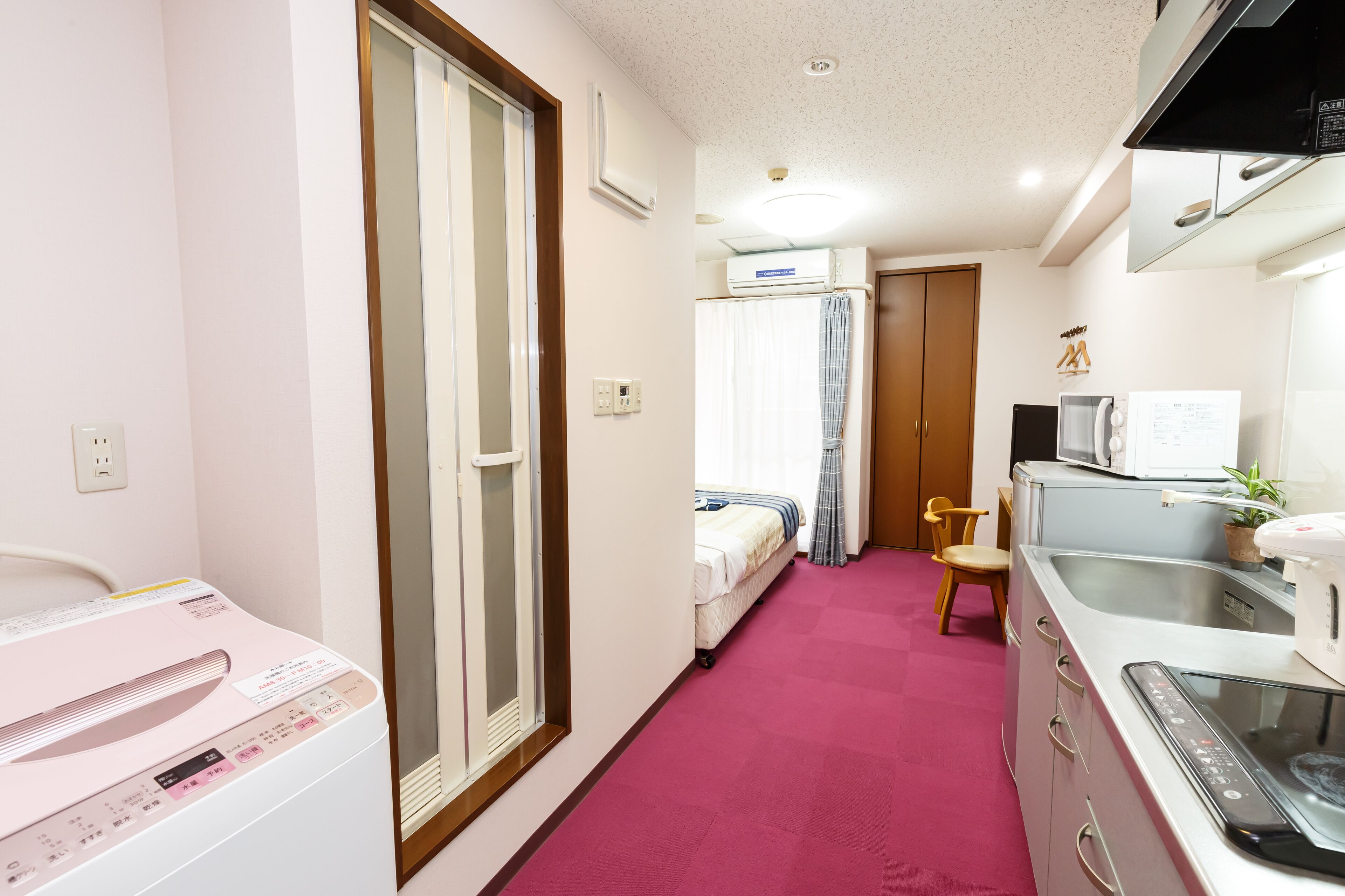 A compact hotel room with a kitchenette, a bed, a small bathroom, and a pink carpet.