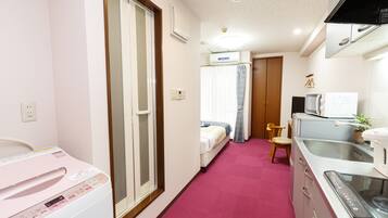 A compact hotel room with a kitchenette, a bed, a small bathroom, and a pink carpet.