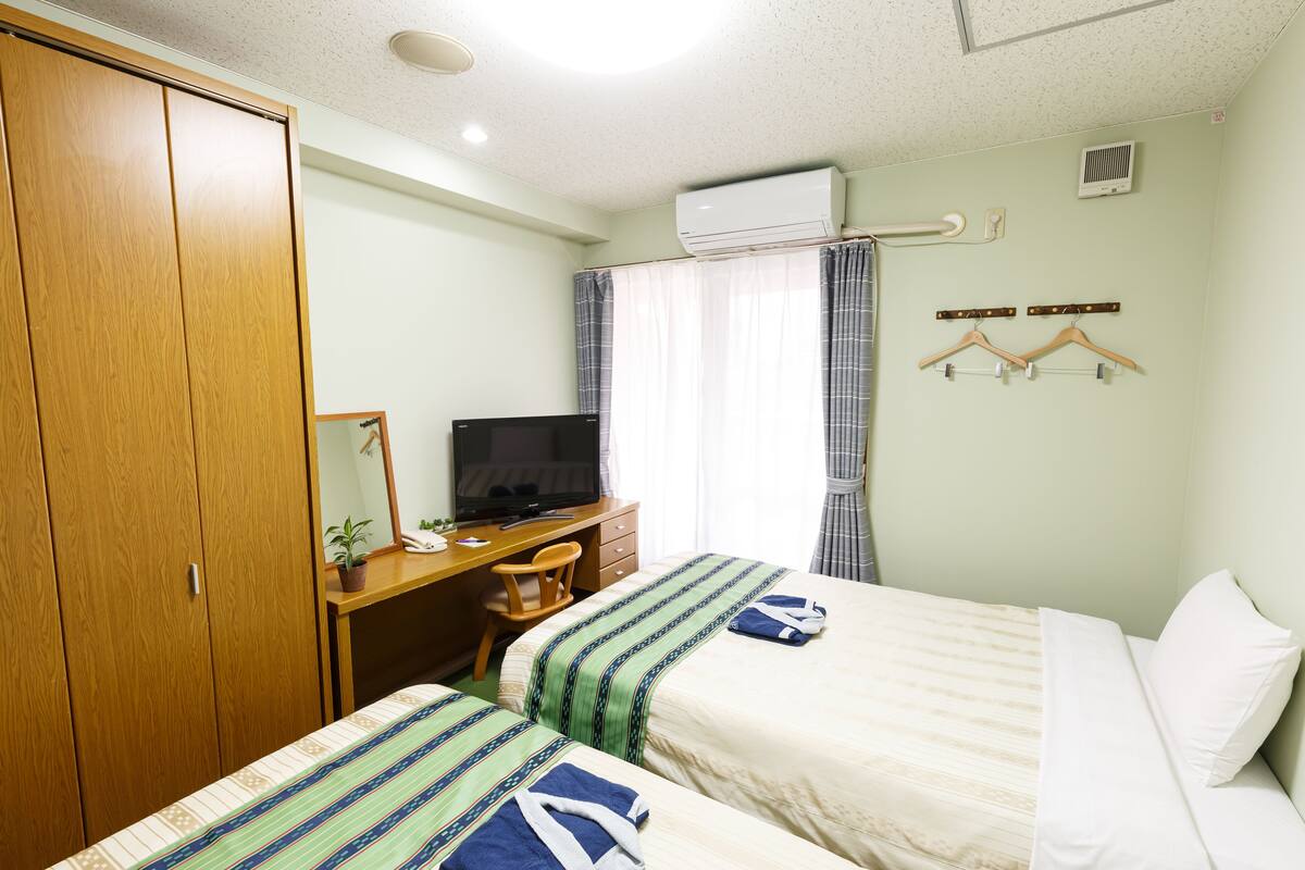 A hotel room with two beds, a desk with a television, a wardrobe, and a window with curtains.