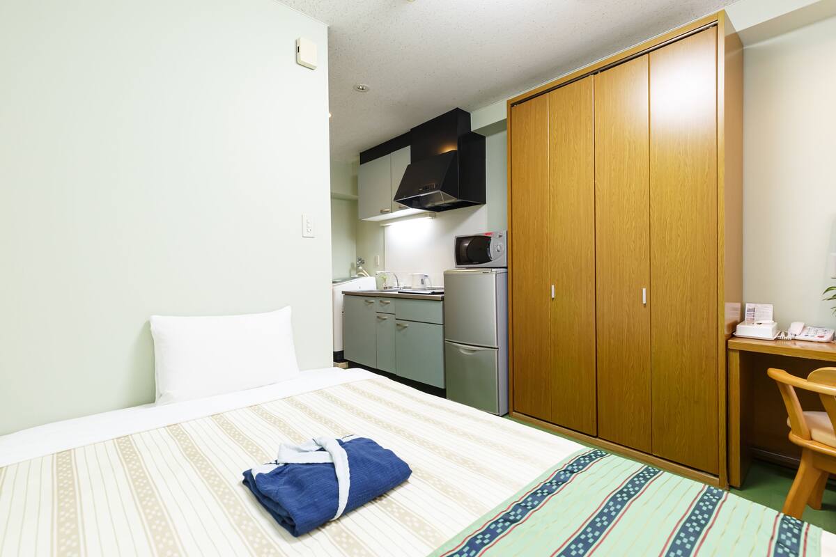 A compact hotel room with a bed, a desk, a kitchenette, and a wardrobe.