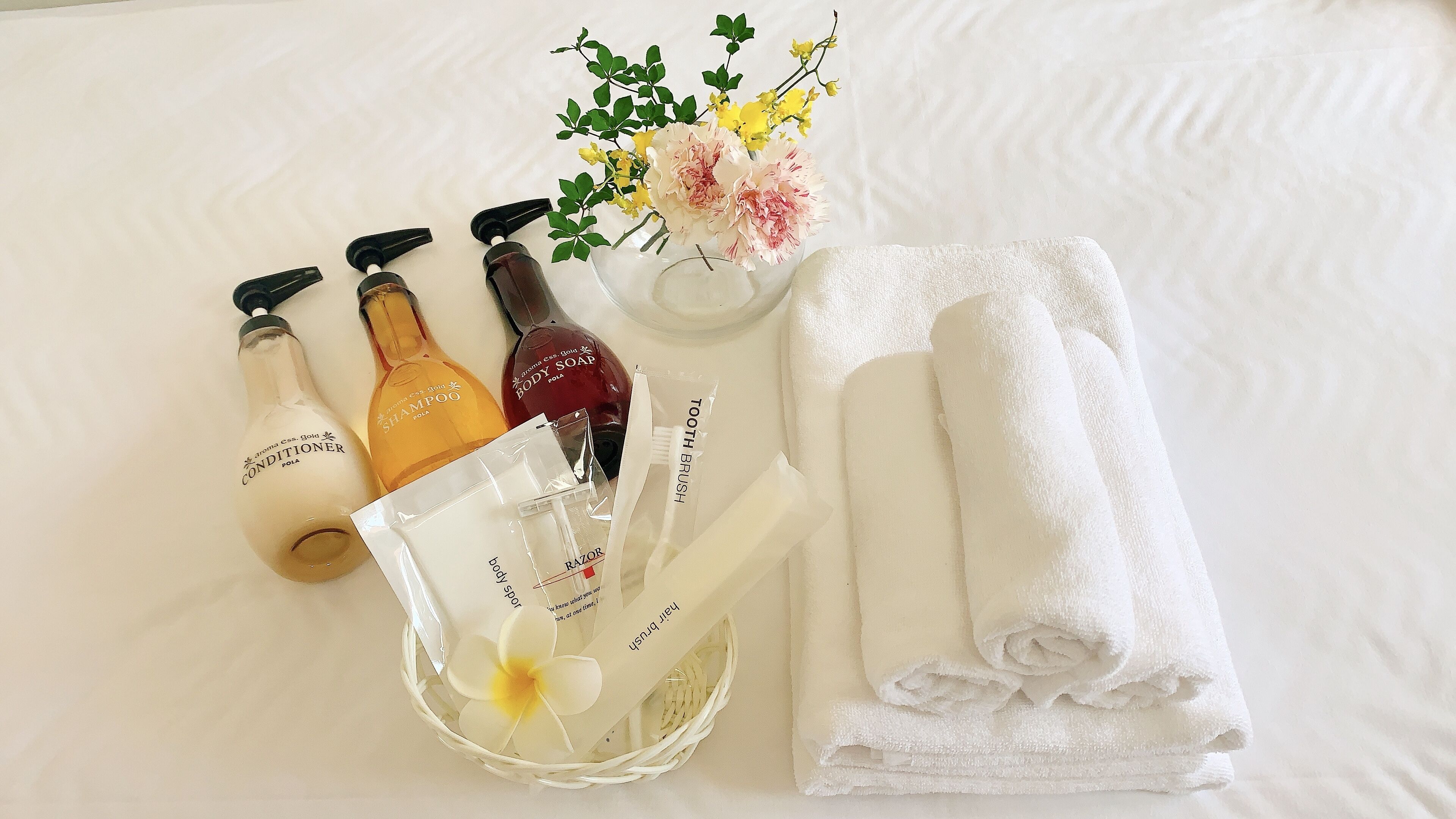 A neatly arranged bathroom with toiletries including shampoo, body soap, and a toothbrush, along with a white towel and a decorative flower arrangement.