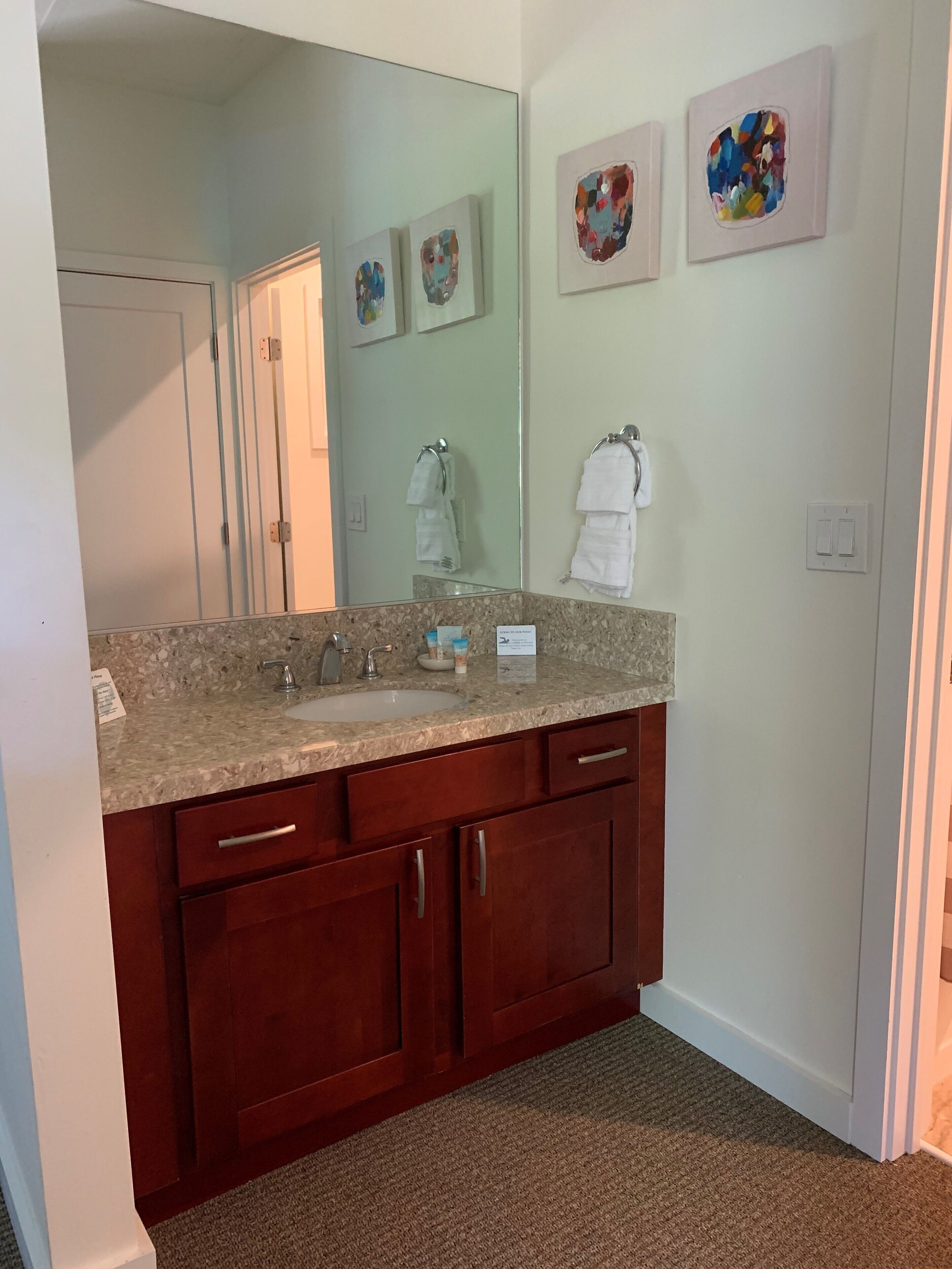 A bathroom with a granite countertop, two sinks, wooden cabinets, a large mirror, and three abstract art pieces on the wall.