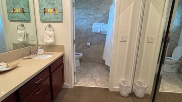A bathroom with a sink, mirror, and two towels hanging on hooks. There are two trash bins and a shower area visible.