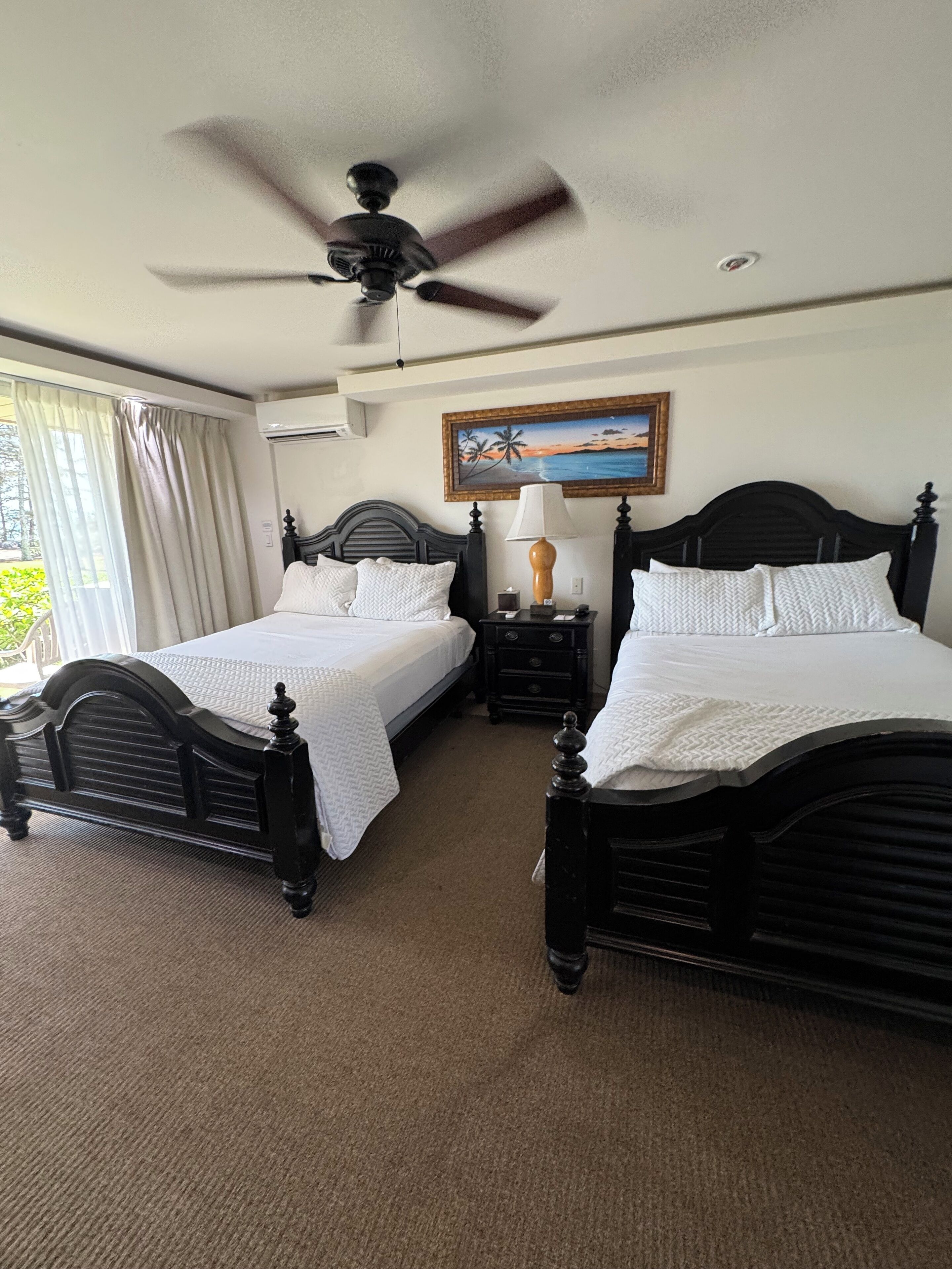 A hotel room with two black beds, a ceiling fan, a painting of a beach scene, and a window with curtains.