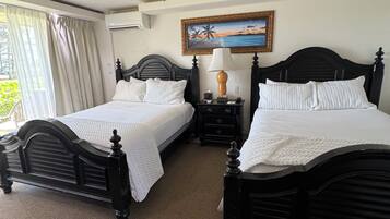 A hotel room with two black beds, a ceiling fan, a painting of a beach scene, and a window with curtains.