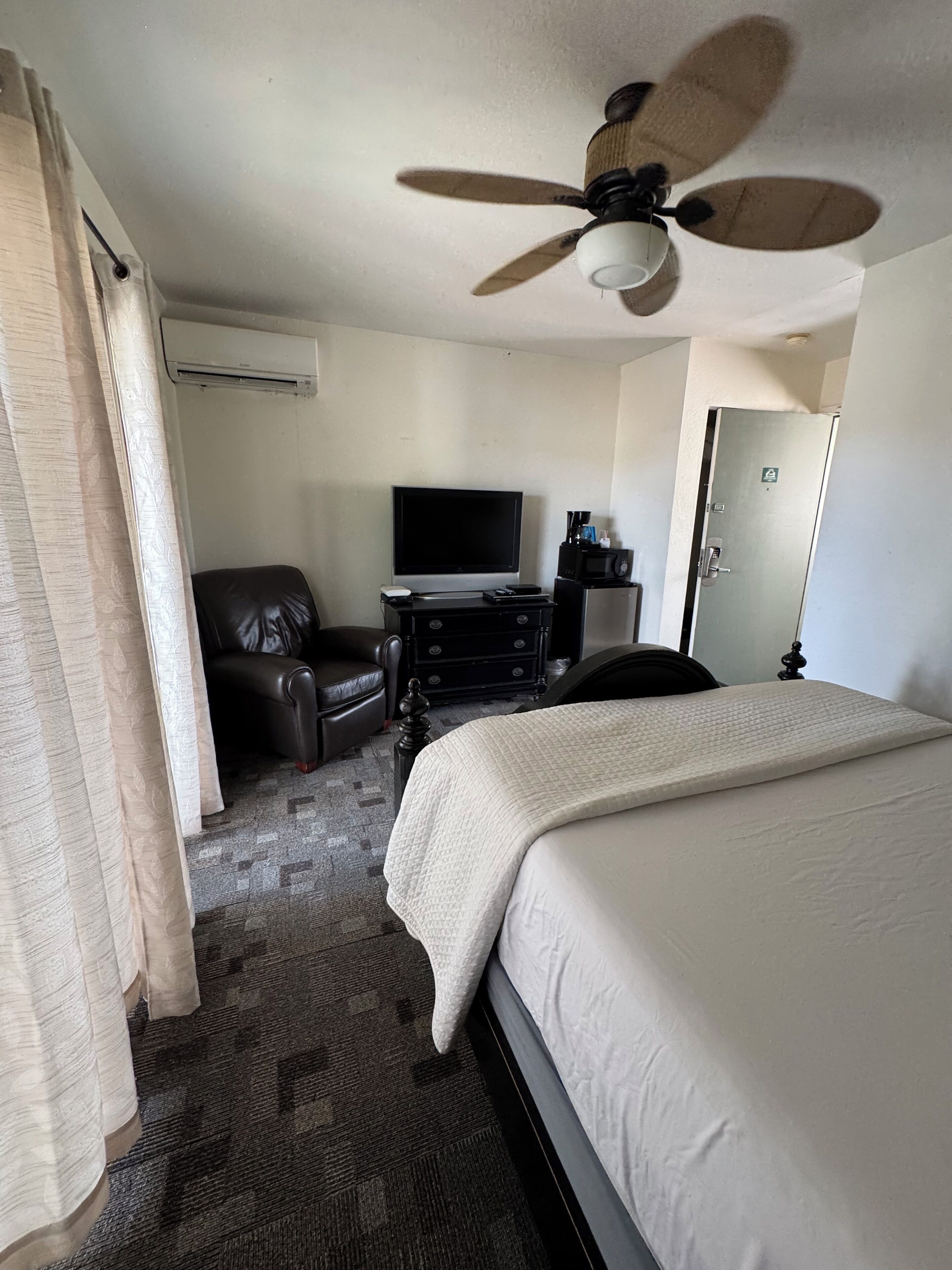A hotel room with a bed, a desk with a computer, a chair, a television, and a ceiling fan.