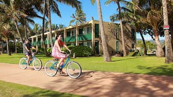 Two people riding bicycles on a sidewalk with palm trees and a building in the background.