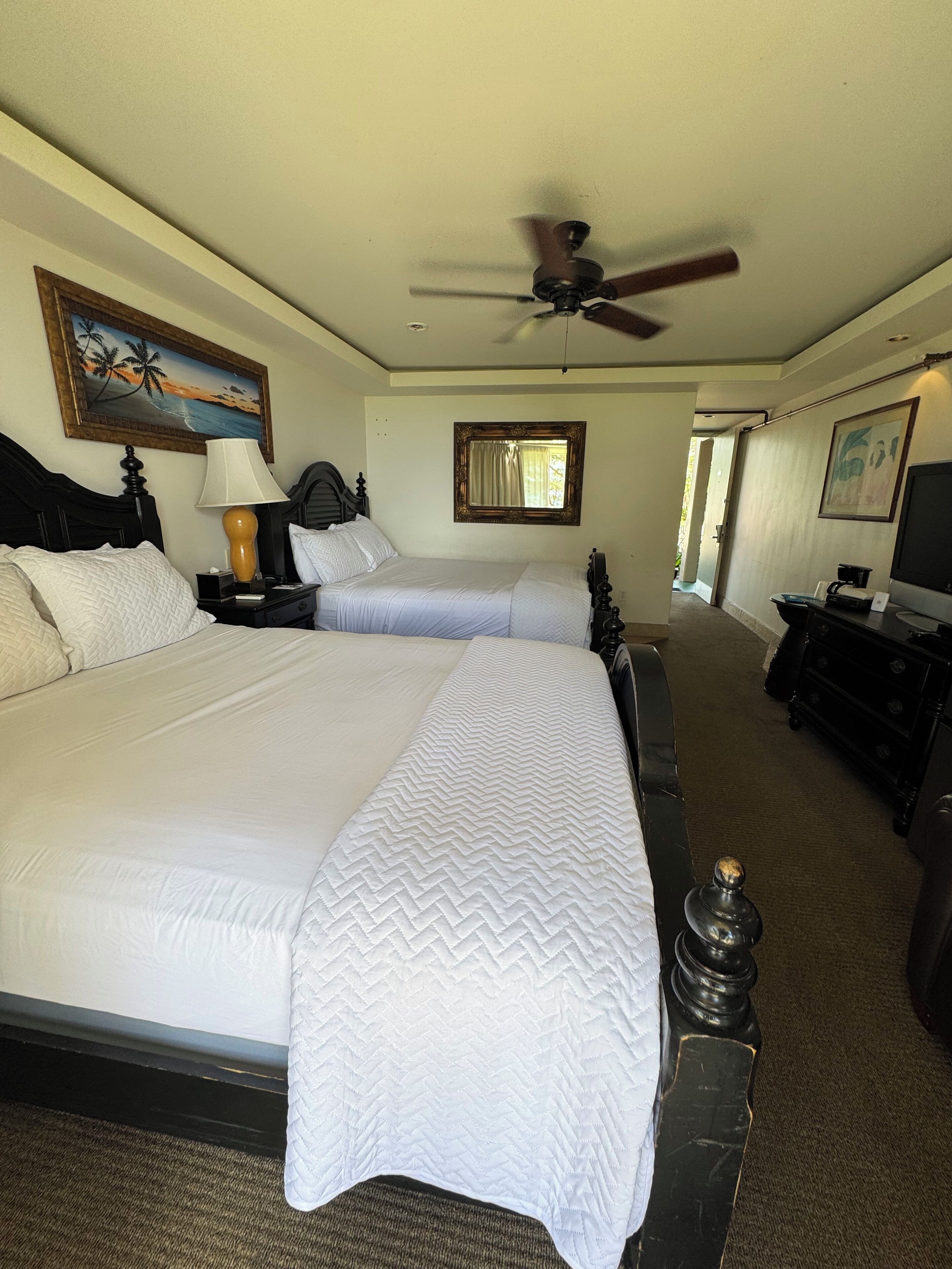 A hotel room with two beds, a ceiling fan, a painting, a lamp, and a dresser.
