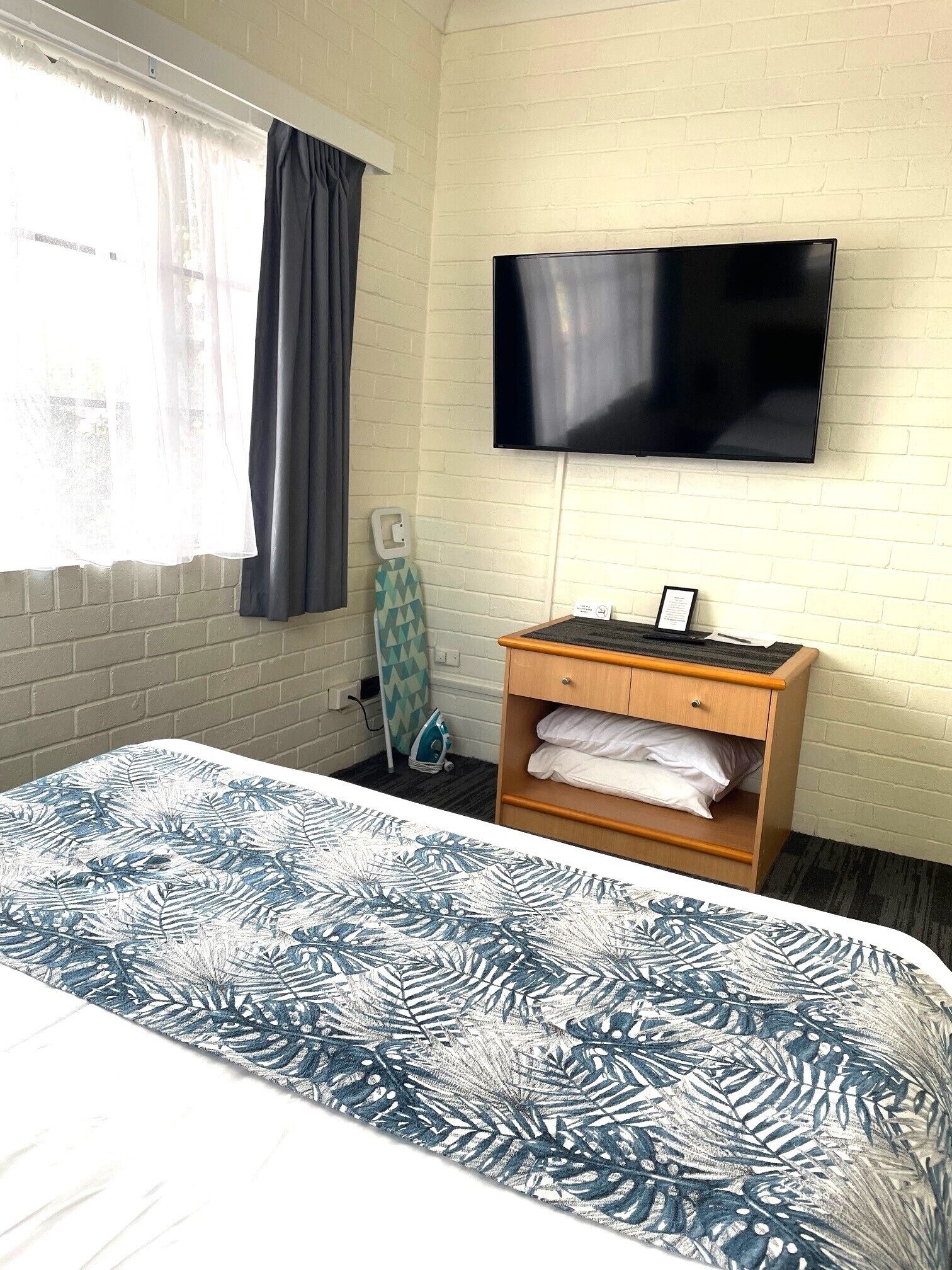 Hotelkamer met een bed, een flatscreen televisie aan de muur, een houten nachtkastje met een lamp en een raam met gordijnen.