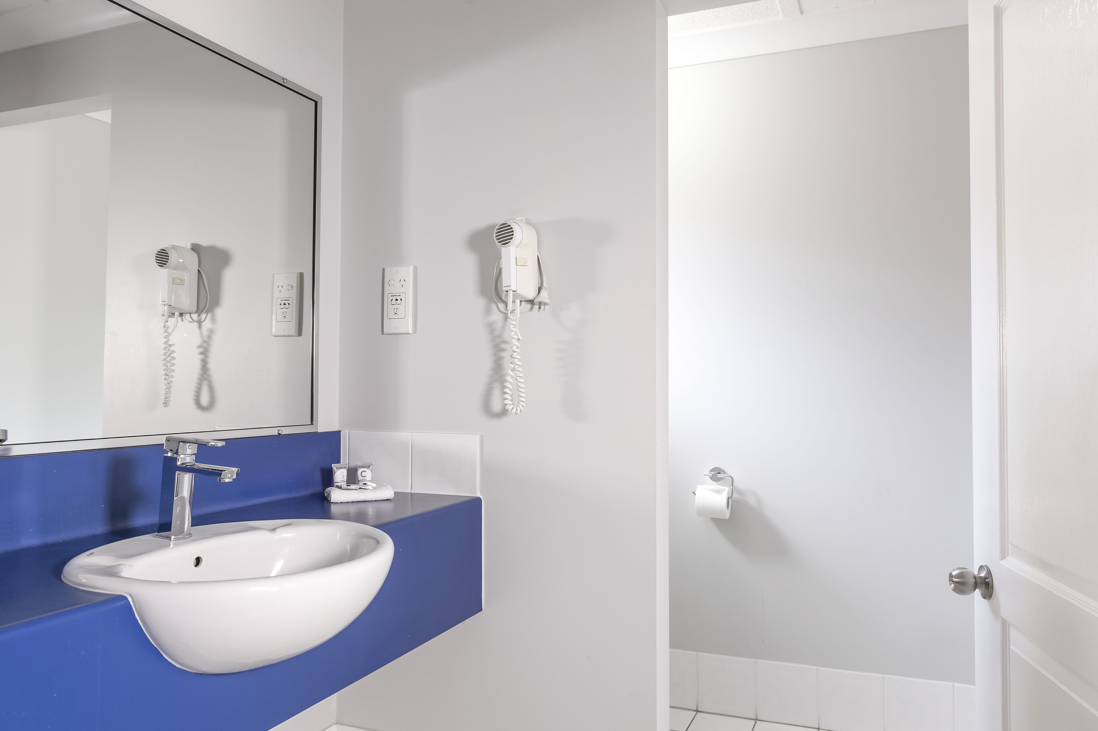 Casa de banho moderna com lavatório branco, bancada azul e dois secadores de cabelo fixados na parede.