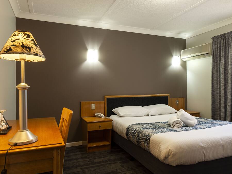 A hotel room with a bed, bedside table, lamp, and a wall-mounted air conditioner.