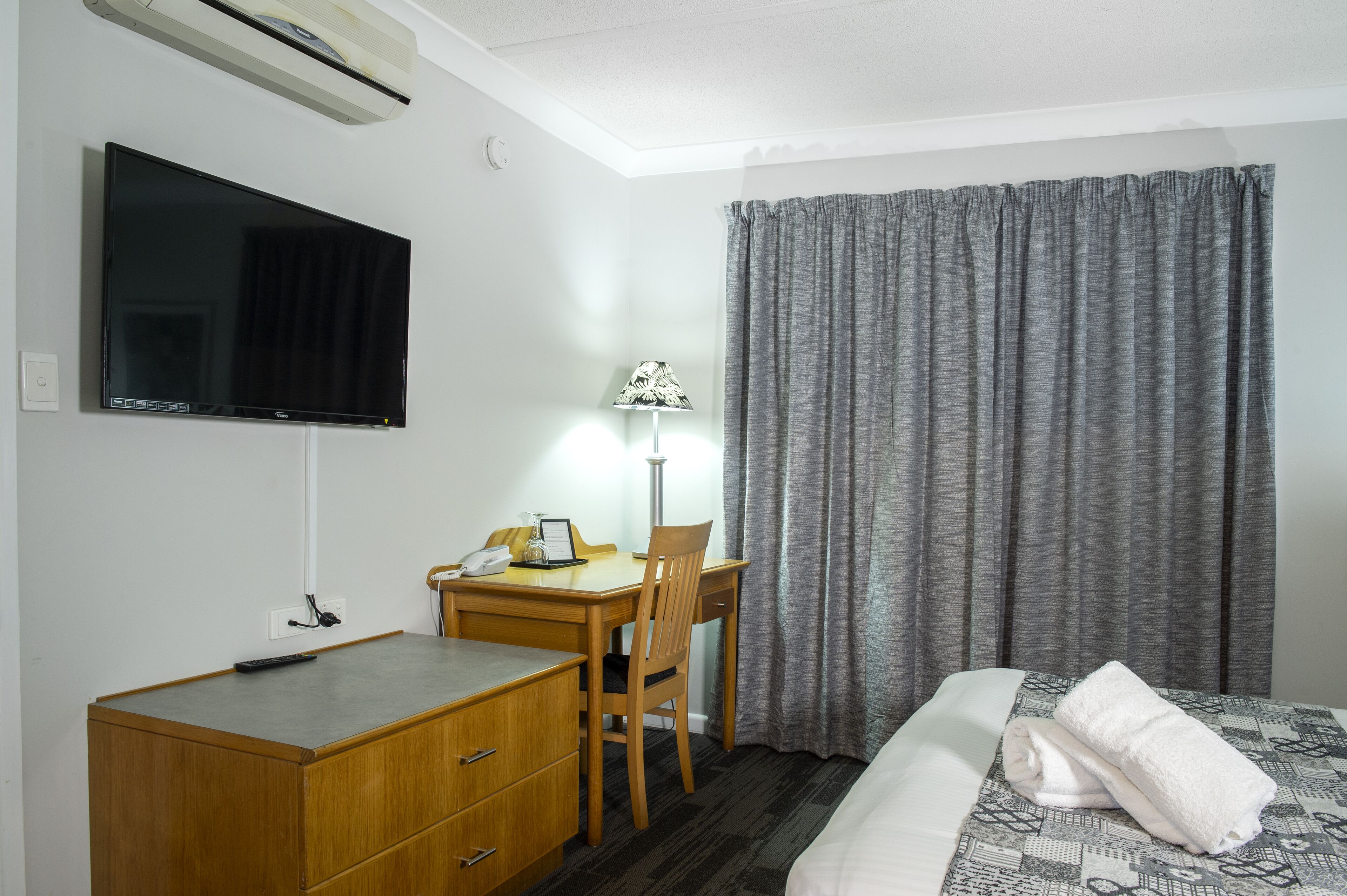 Una habitación de hotel con una cama, un escritorio con una silla, un televisor montado en la pared y una ventana con cortinas.
