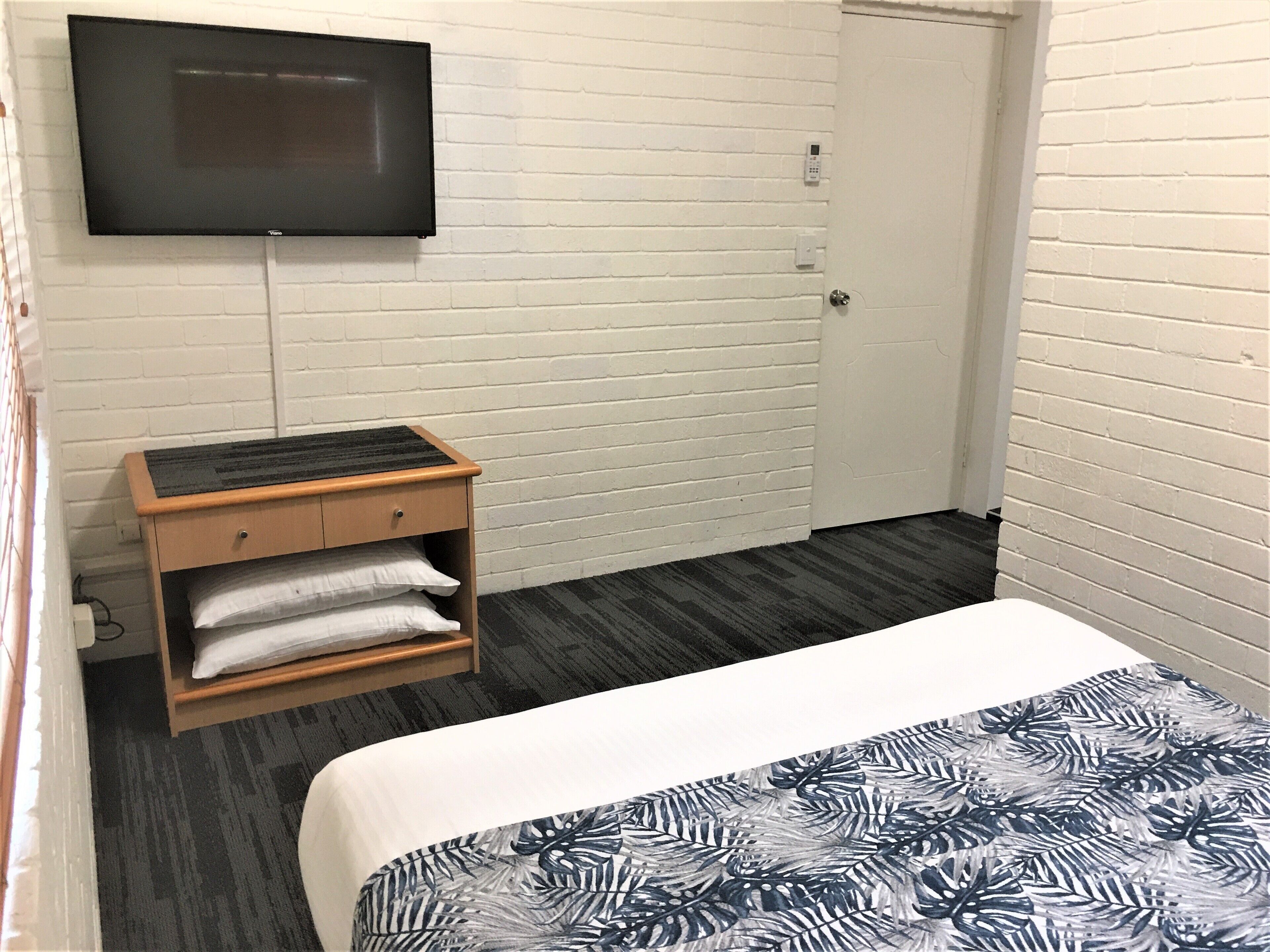 Una habitación de hotel con una cama, un televisor montado en la pared, una mesita de noche de madera con una lámpara y una puerta.