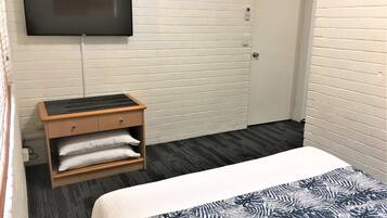 Una habitación de hotel con una cama, un televisor montado en la pared, una mesita de noche de madera con una lámpara y una puerta.