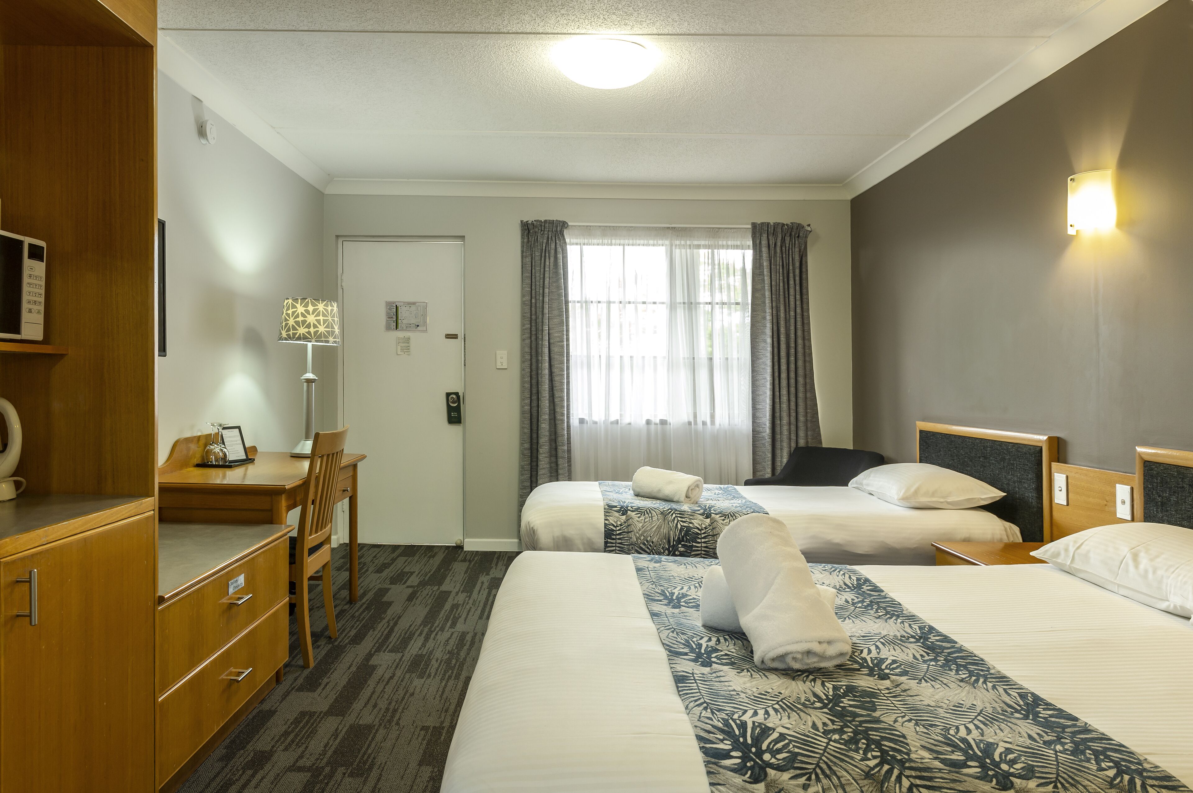 Habitación de hotel con dos camas, un escritorio con silla, una lámpara y un armario.