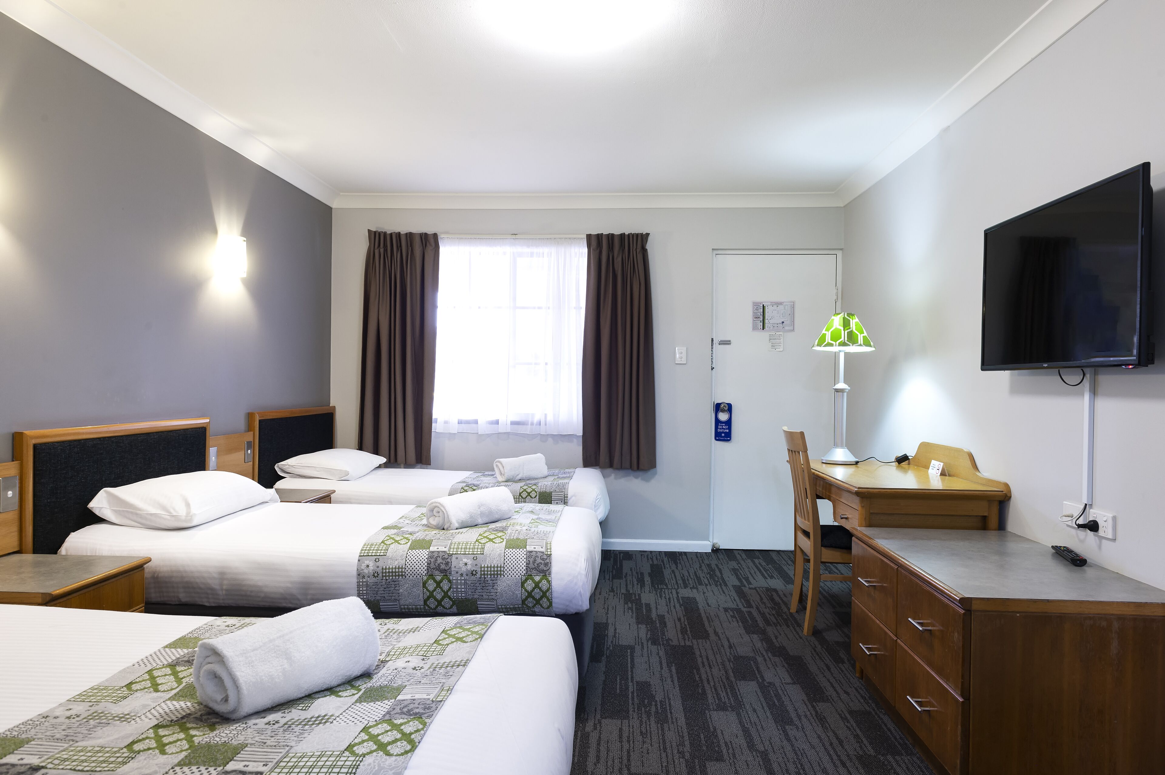 A hotel room with three beds, a desk, a chair, a television, and a window with curtains.