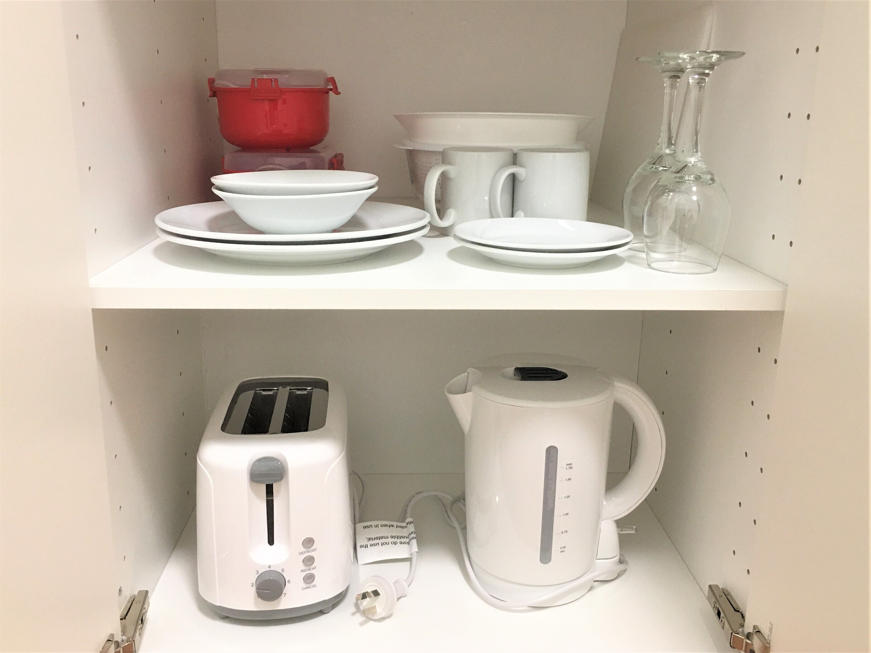 Uma prateleira na cozinha com pratos brancos, tigelas, um recipiente vermelho e uma chaleira branca.