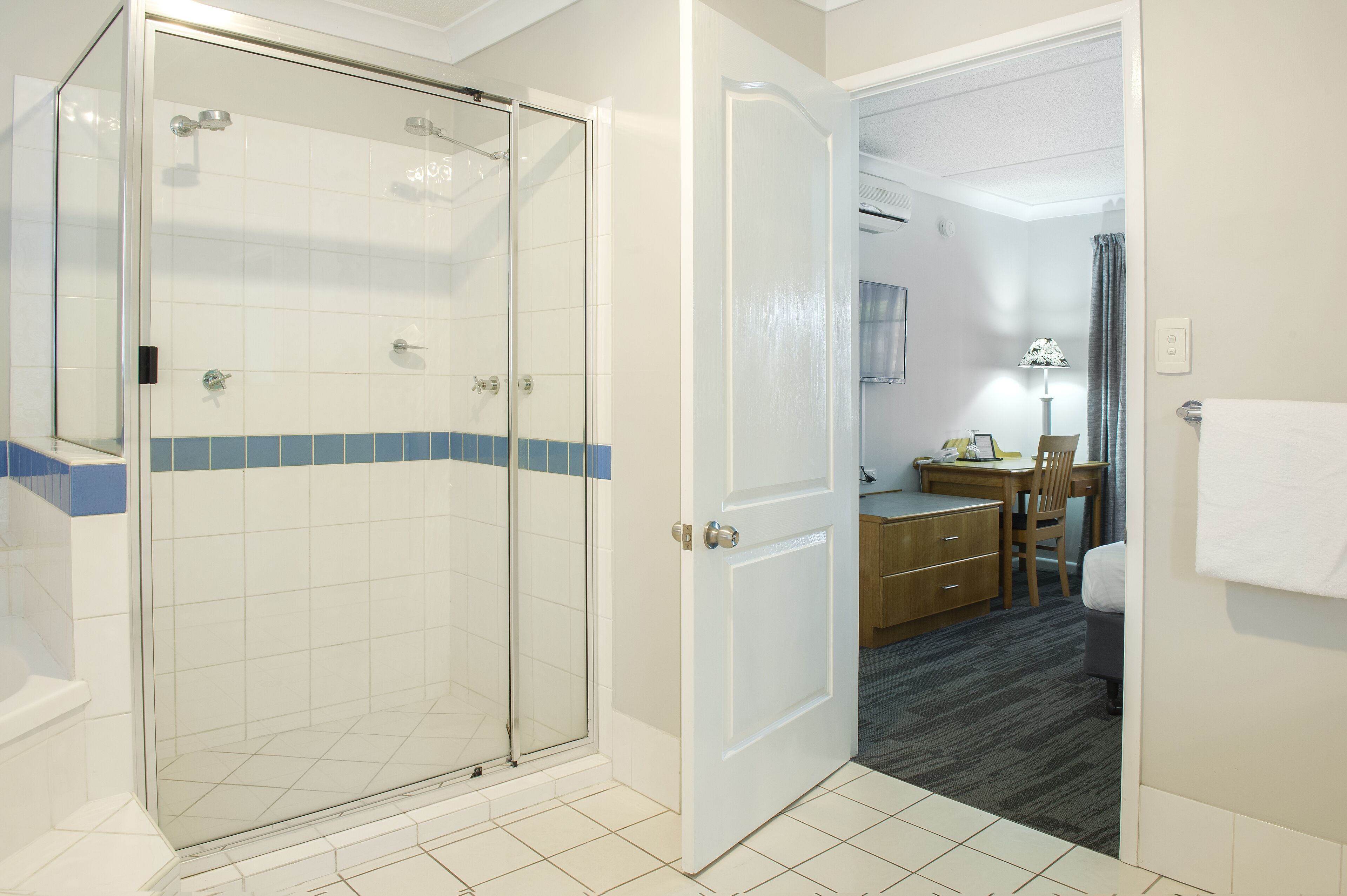 Un baño de hotel con ducha envuelta en vidrio, una puerta blanca y un lavabo visible sobre el mueble.