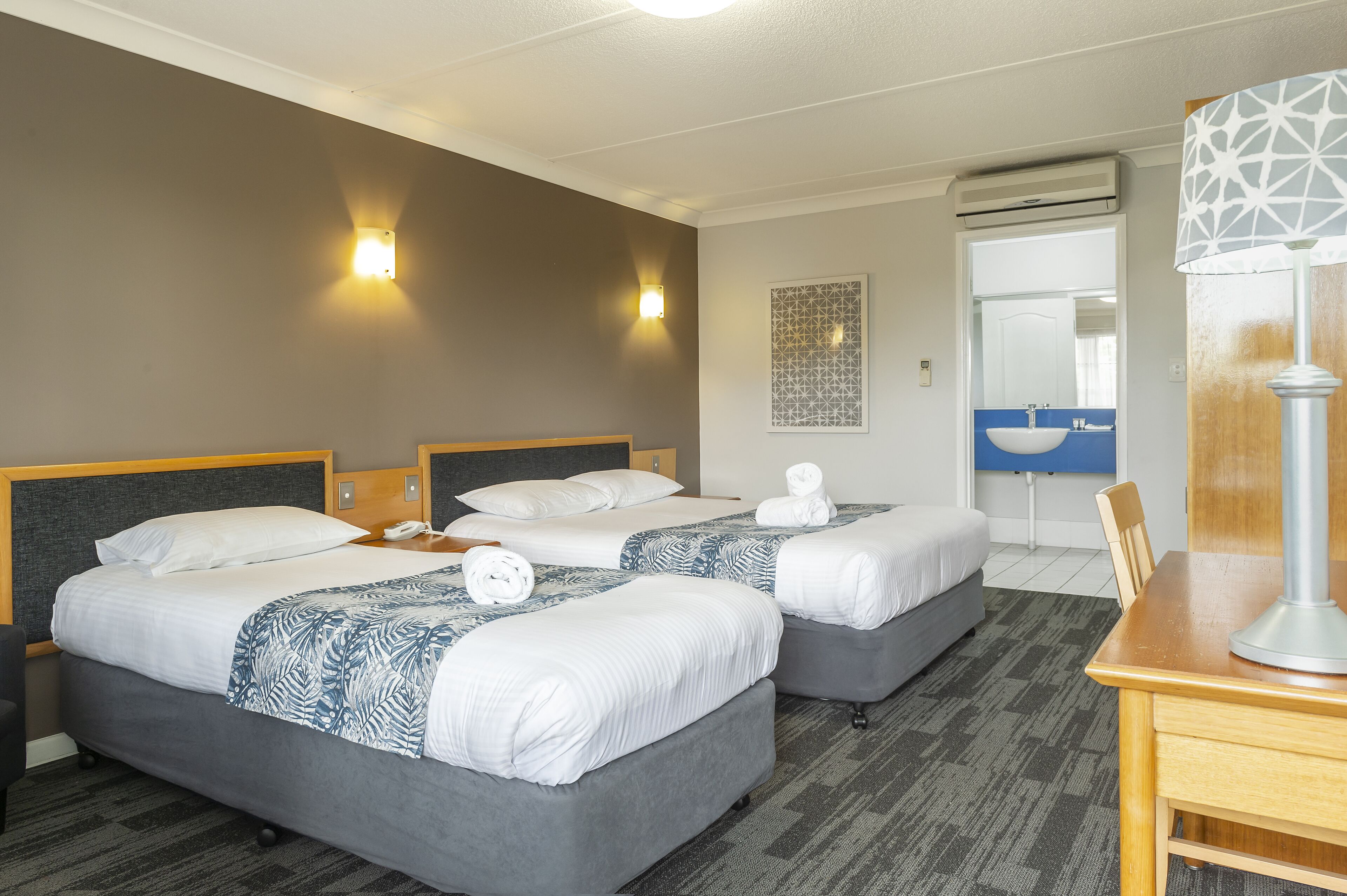 A hotel room with two beds, a desk, a lamp, and a bathroom visible through an open door.