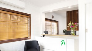 Un área de recepción con un mostrador blanco, dos sillas negras, persianas de madera, un reloj, un jarrón con flores y una computadora Lenovo.