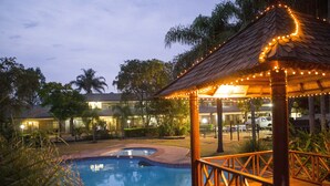 Un área de piscina con cubierta de madera y pabellón de techo de paja, rodeada de vegetación y edificios.