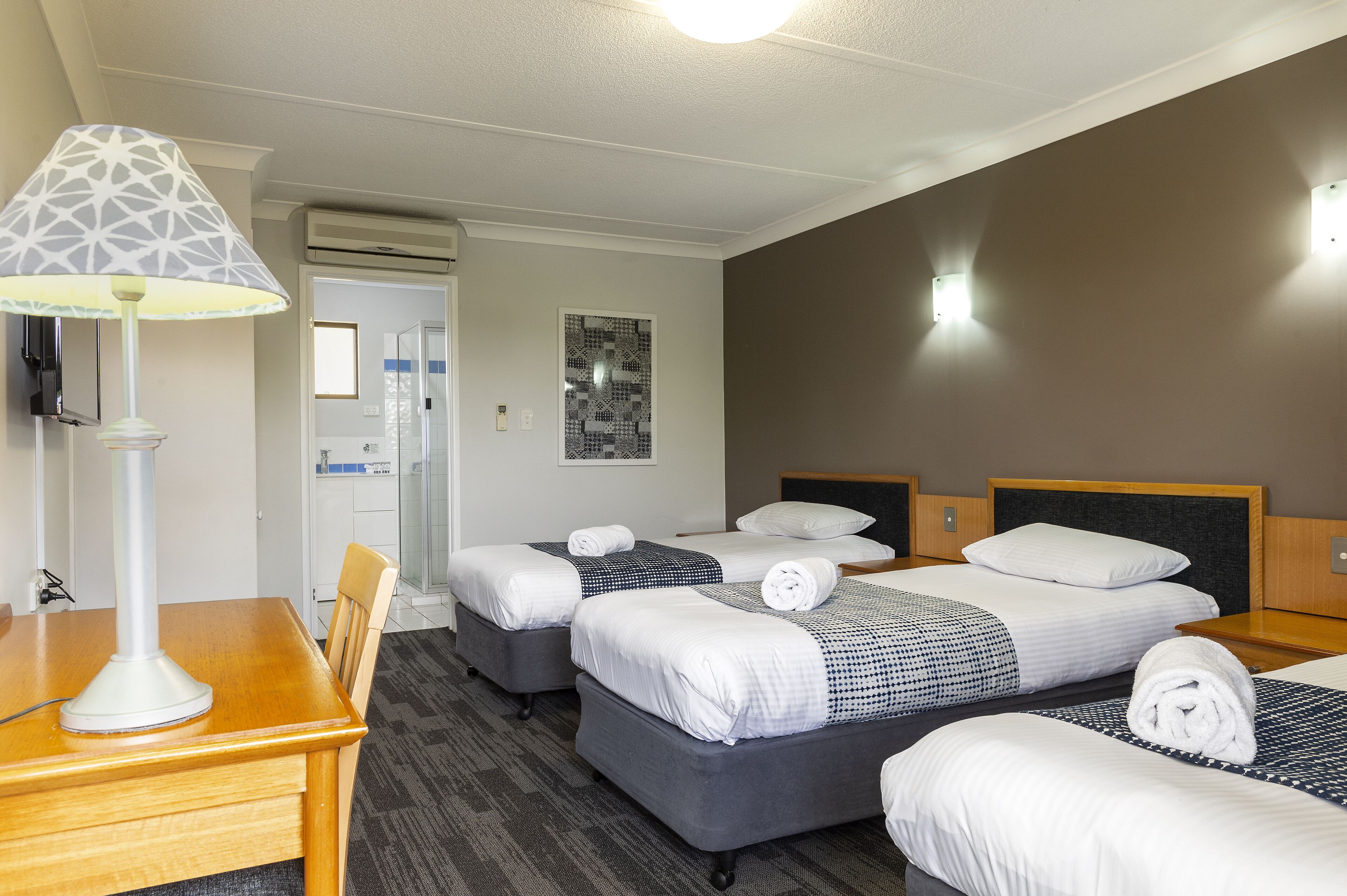 A hotel room with three beds, a desk, a lamp, and an air conditioning unit.