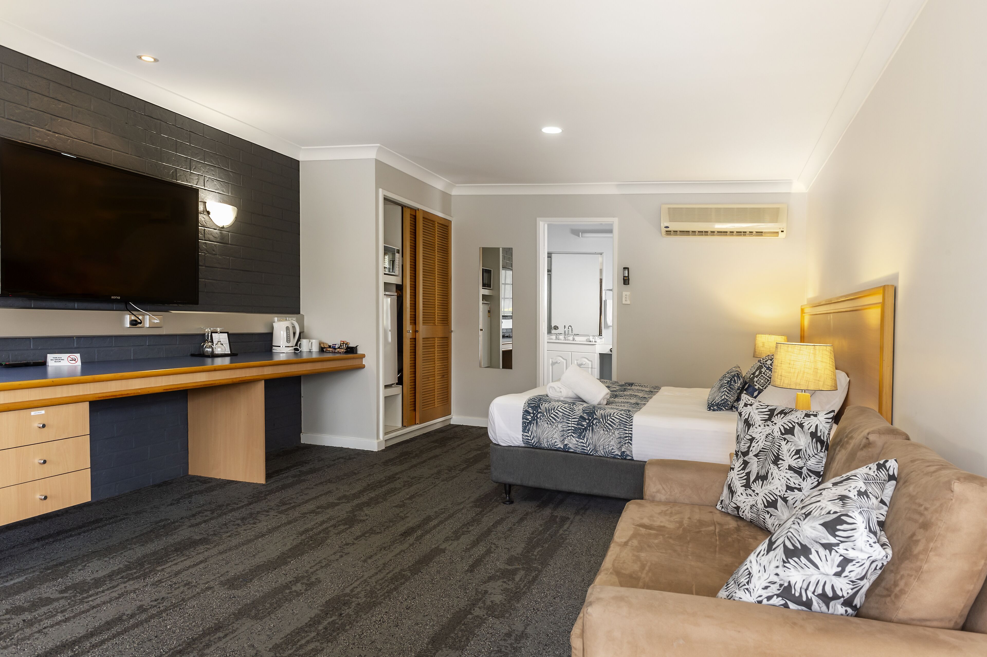 A modern hotel room with a kitchenette, a large flat-screen TV, a bed with patterned pillows, and a sofa.