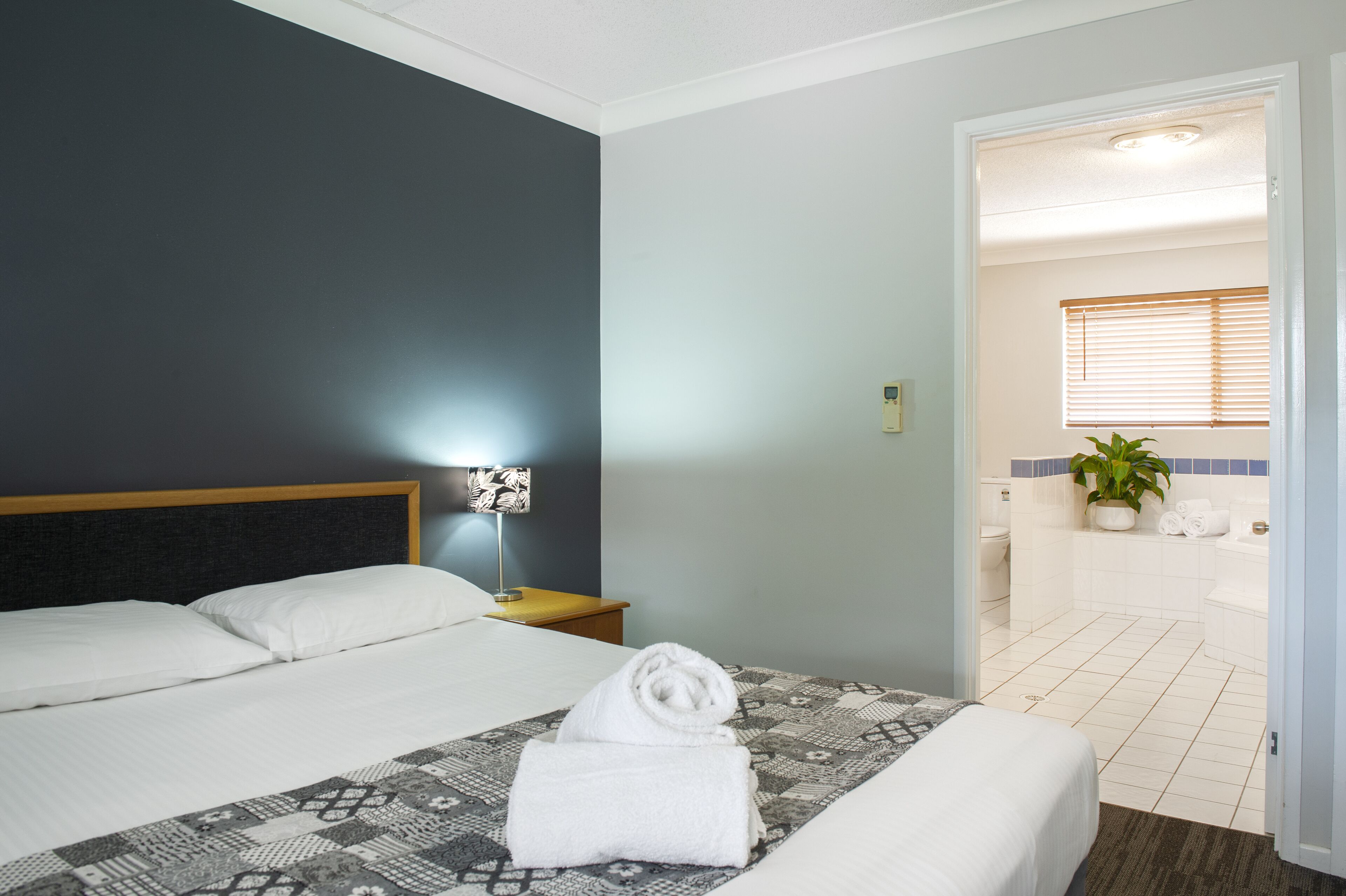 Habitación de hotel con cama, mesita de noche, lámpara, toalla y baño visible a través de una puerta abierta.