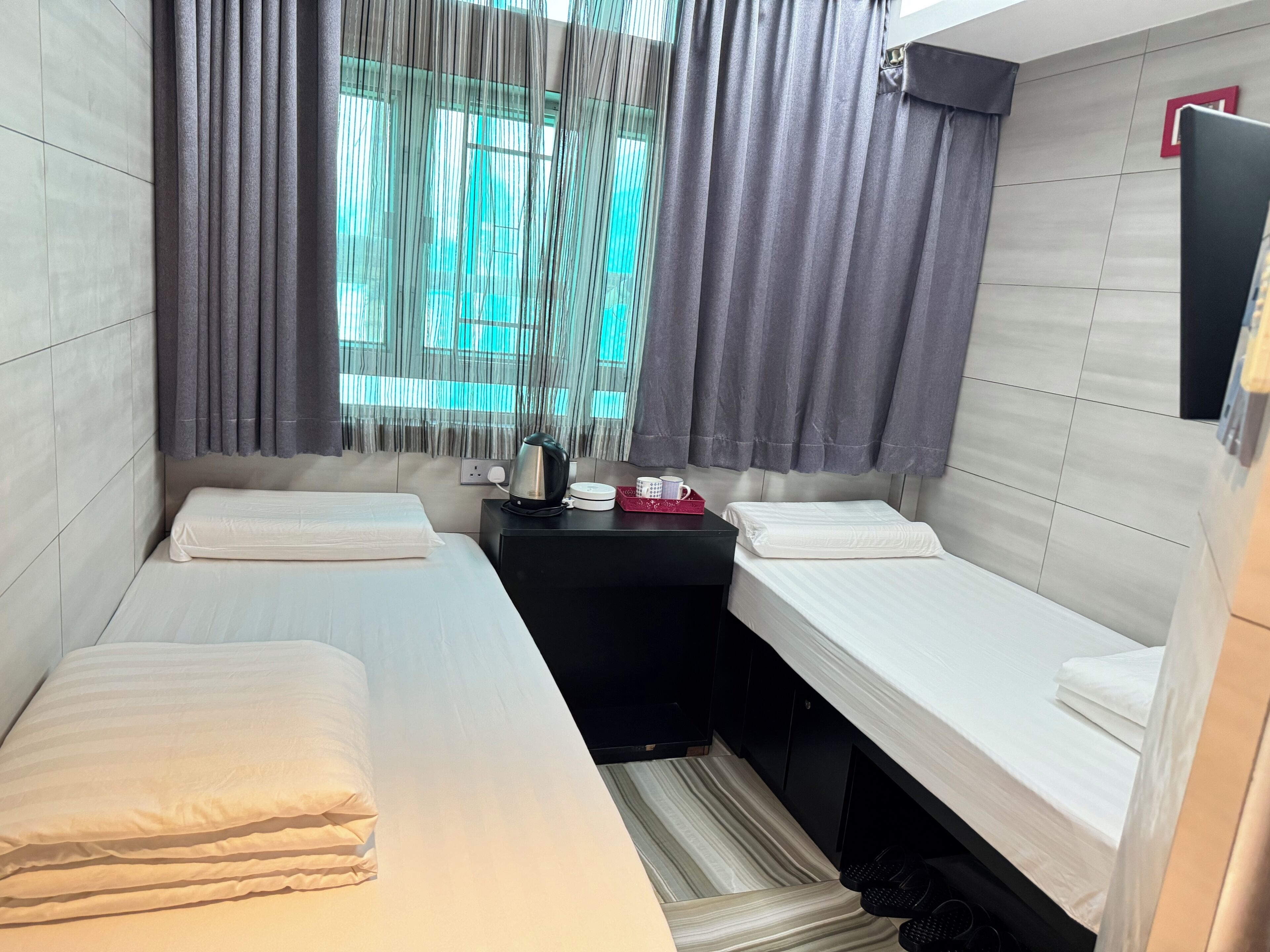 A compact hotel room with two single beds, a small table with a kettle and a mug, and a window with curtains.